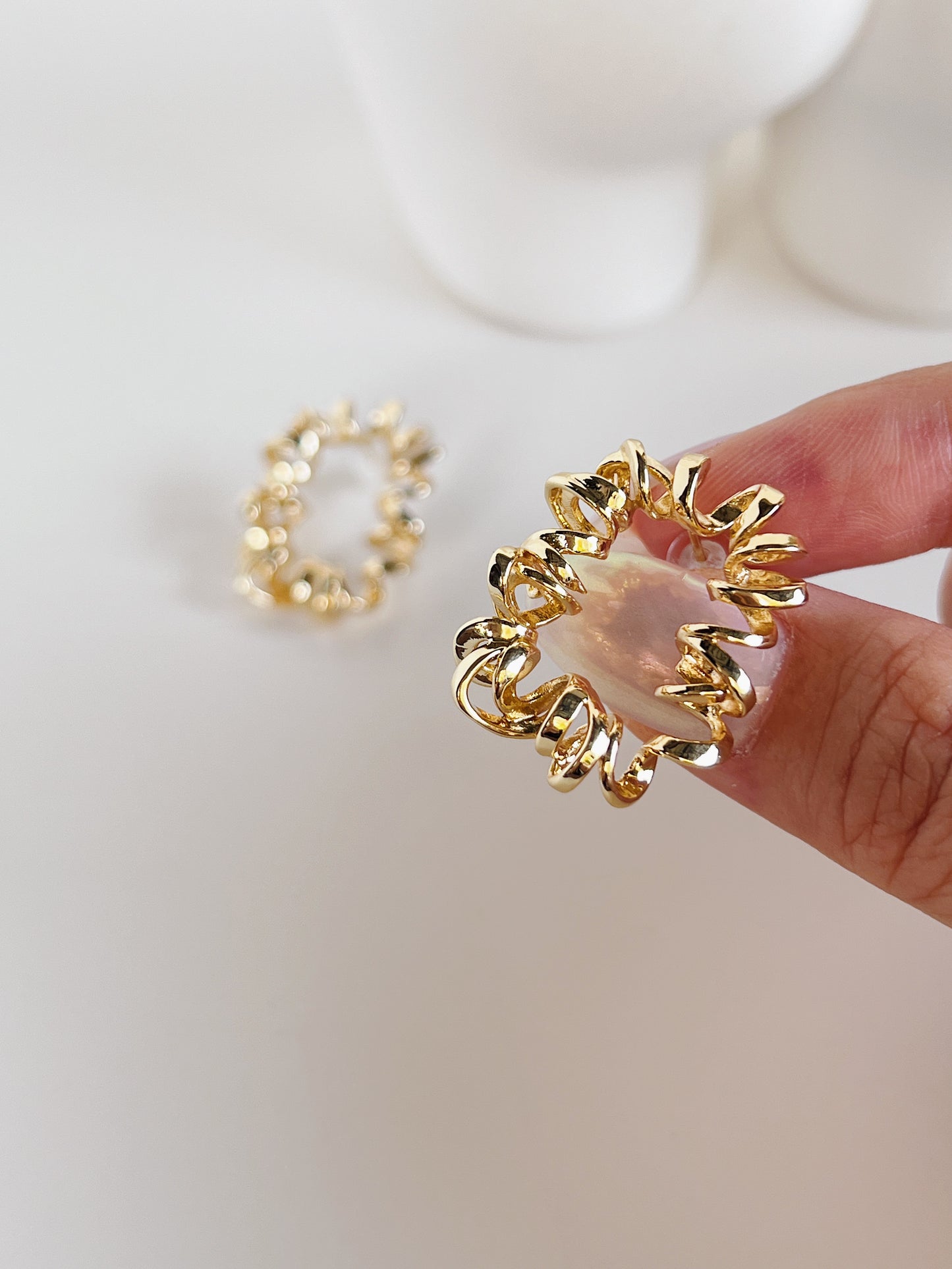 Gold earring with a textured design held by a hand on a white background