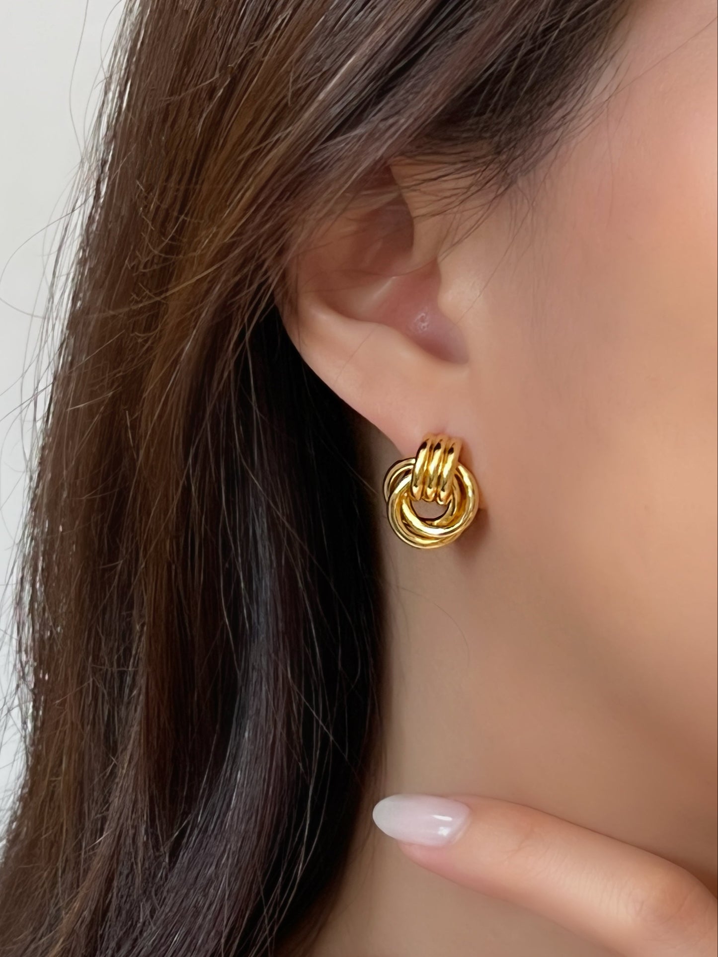 Gold triple knot hoop earrings worn by a person with a neutral background.