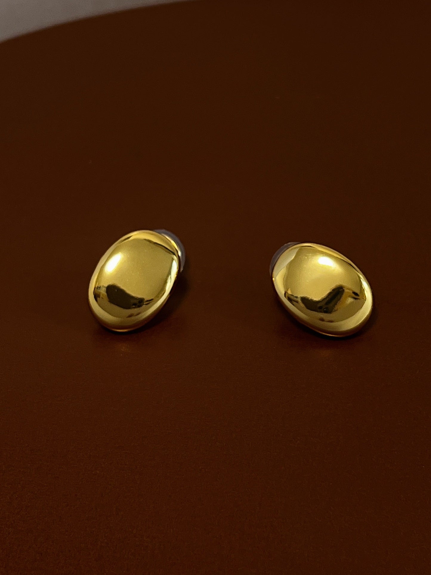 High-polish pair of sleek, gold oval clip on earrings resting on a rich brown leather background.