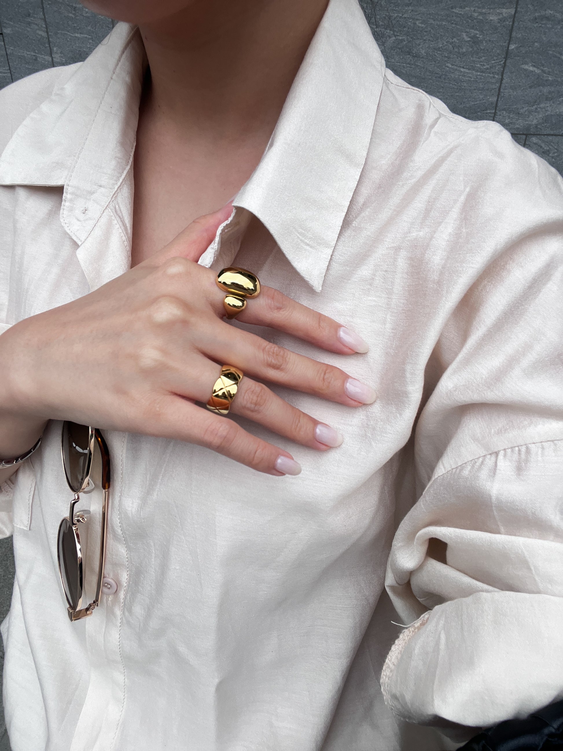 Person wearing gold rings on a beige shirt with sunglasses on the shoulder.