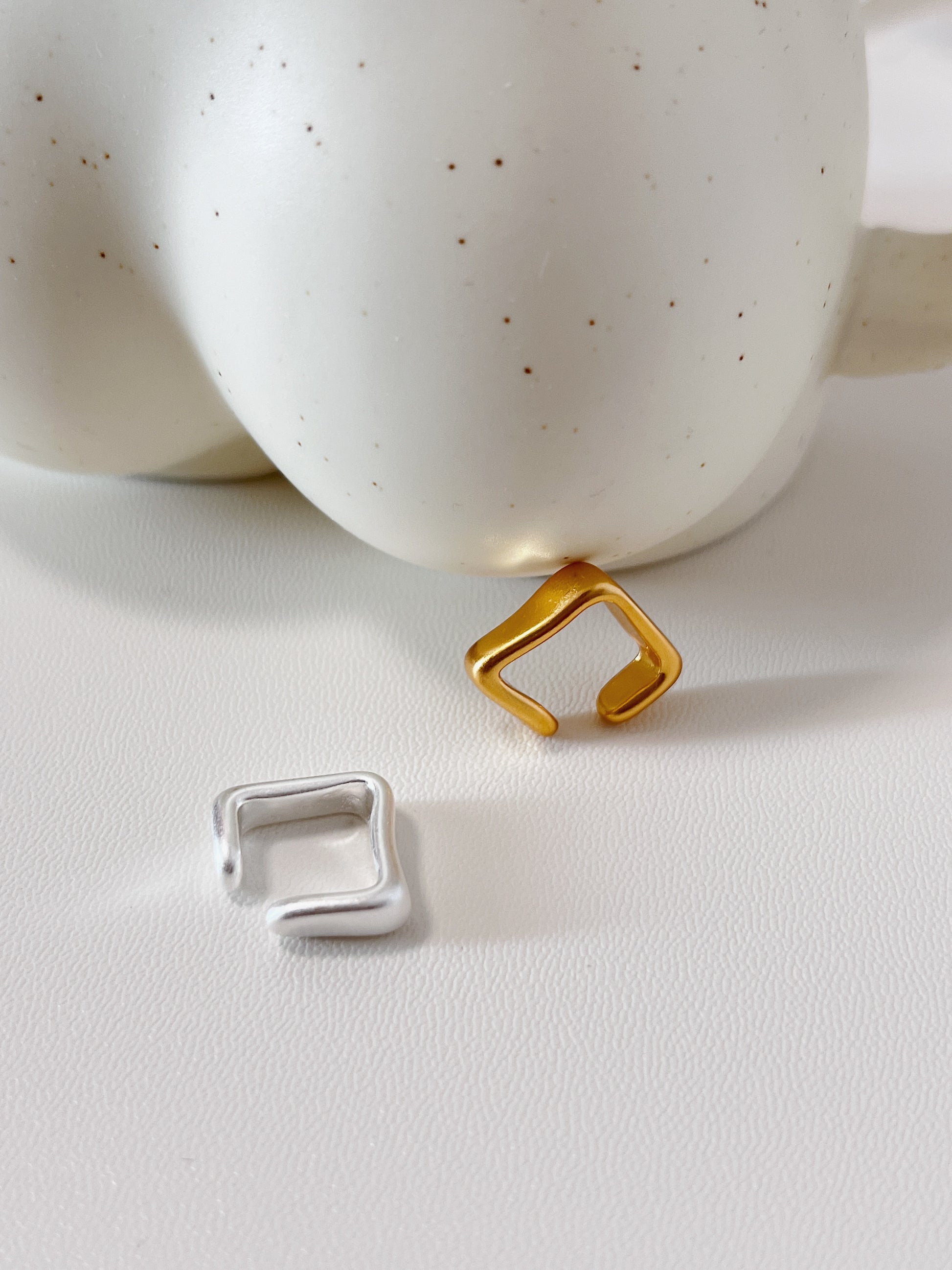 Gold and silver square-shaped ear cuff earrings on a white surface with a ceramic bowl in the background.