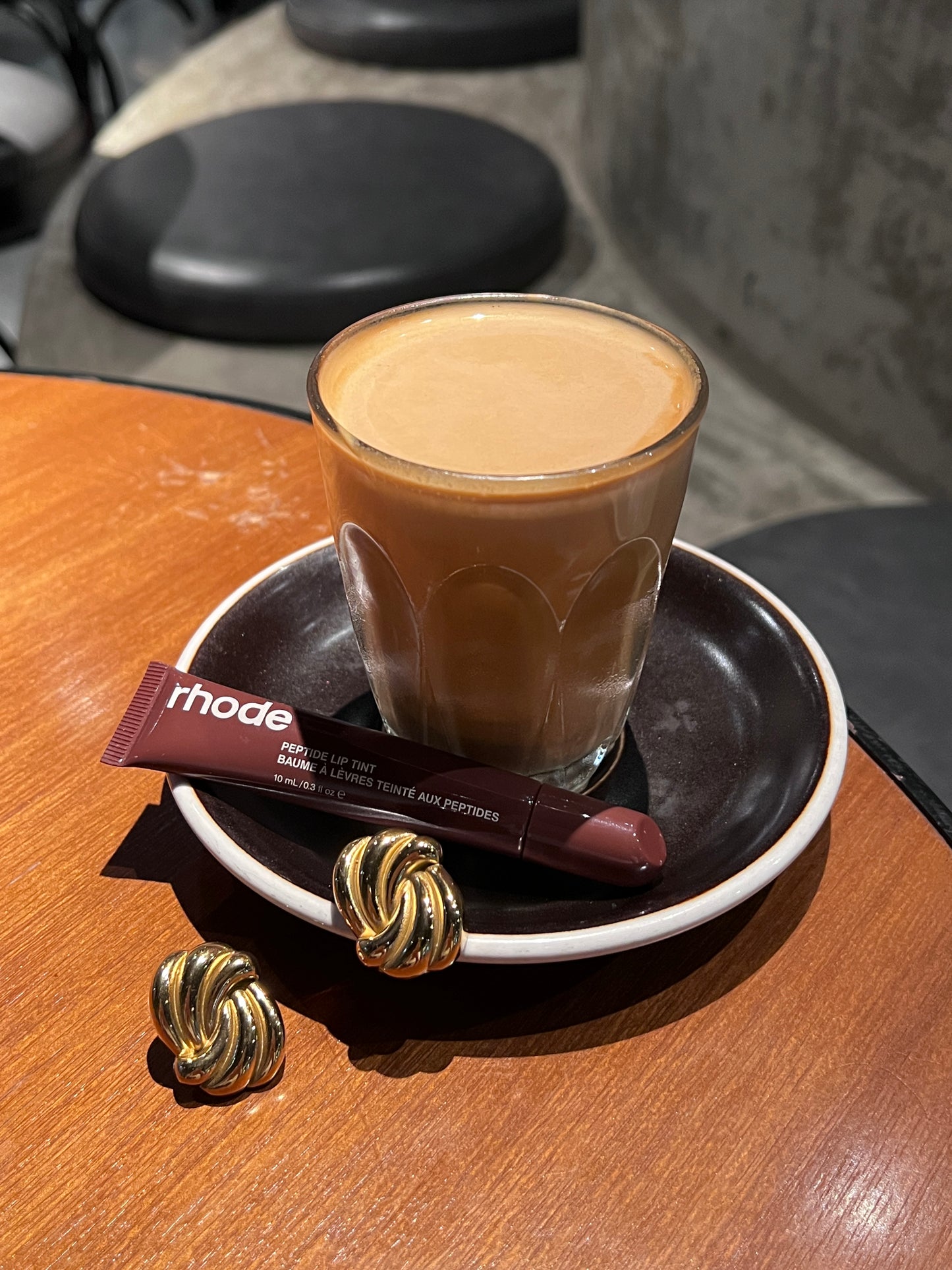 Glass of coffee with a espresso rhode lipgloss and gold stud earrings on a wooden table.