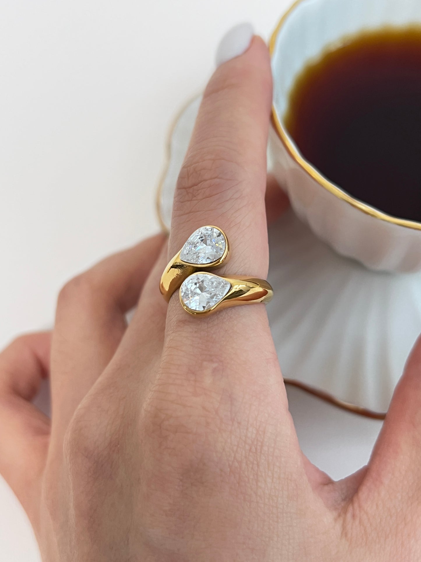 Gold ring with teardrop-shaped gemstones worn on a finger, with a cup of coffee in the background.