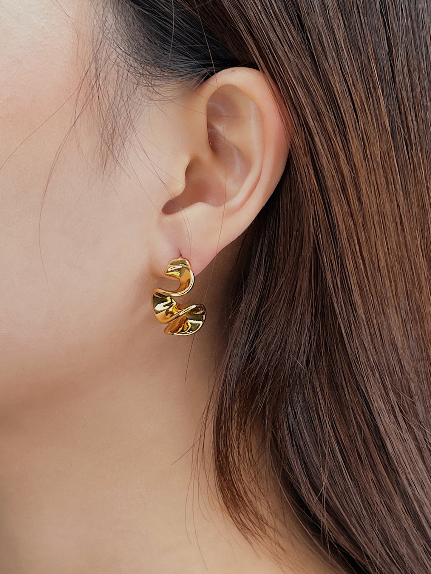 Close-up of a person wearing gold sculptured earrings with a neutral background