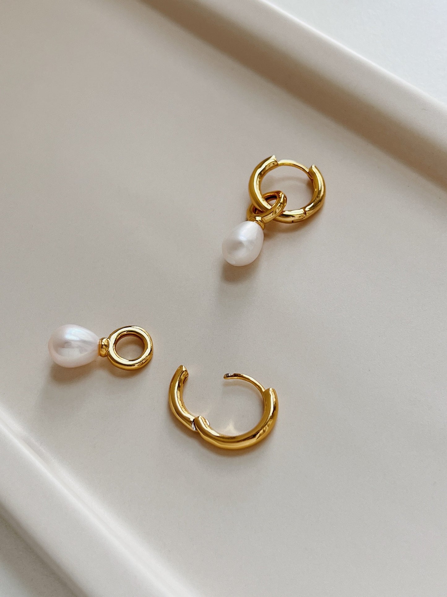 A pair of gold dangle pearl earrings, one placing on top of a white table and another one placing on top of a beige tray plate.