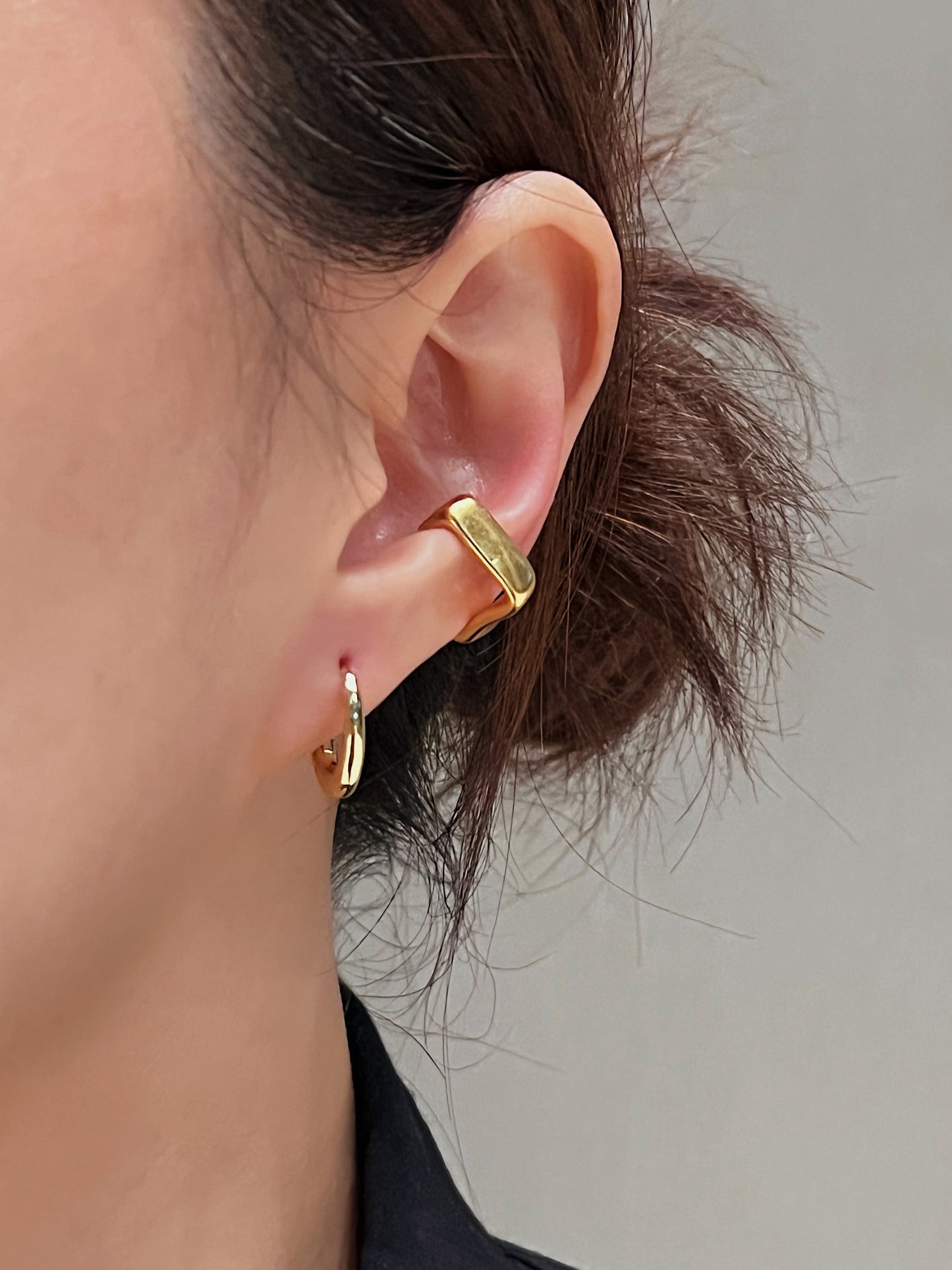 Close-up of a person wearing gold oval hoop earrings and gold brushed ear cuff against a neutral background