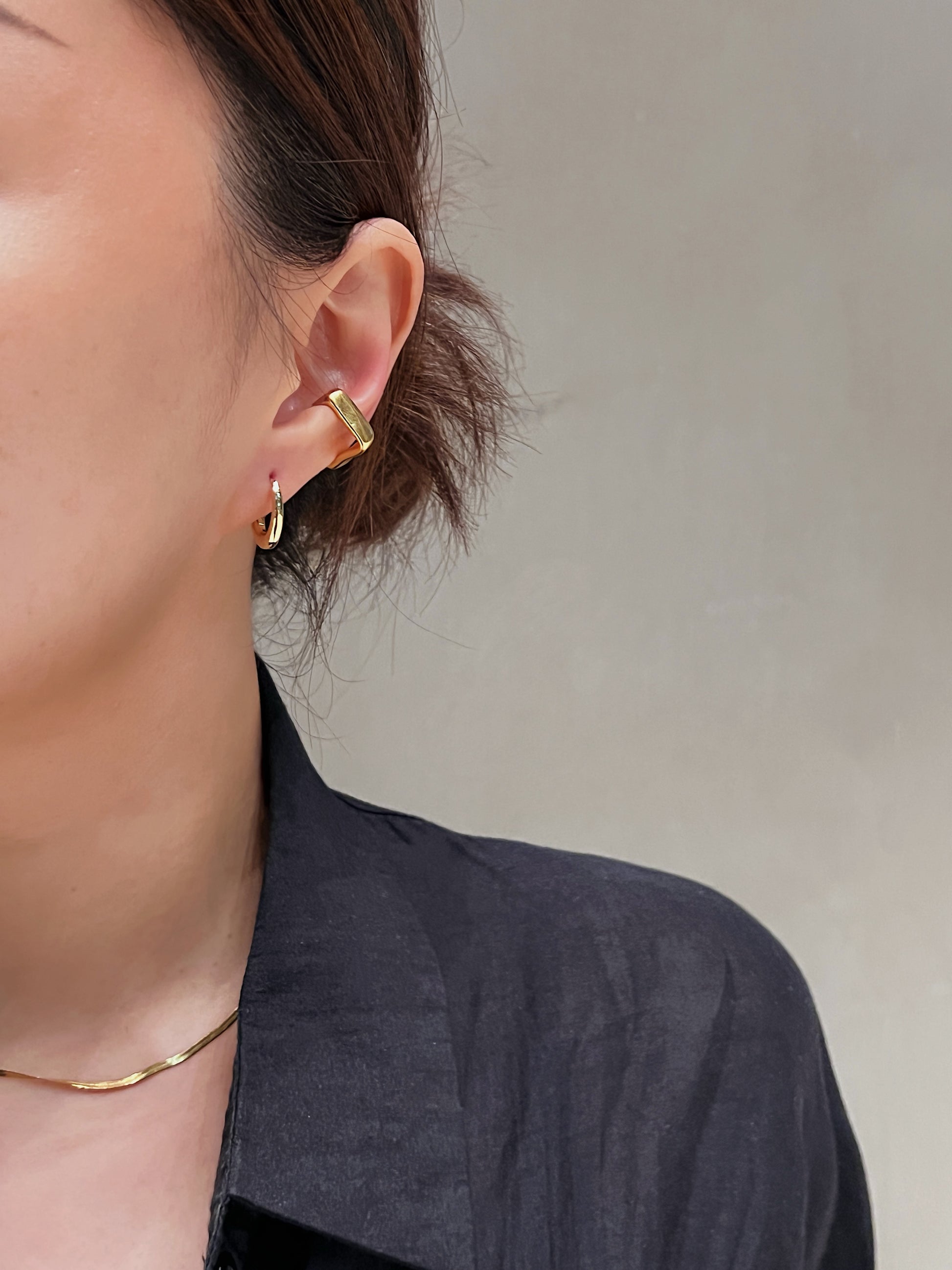 Close-up of a person wearing gold huggie hoop earrings and gold square shape ear cuff with a neutral background