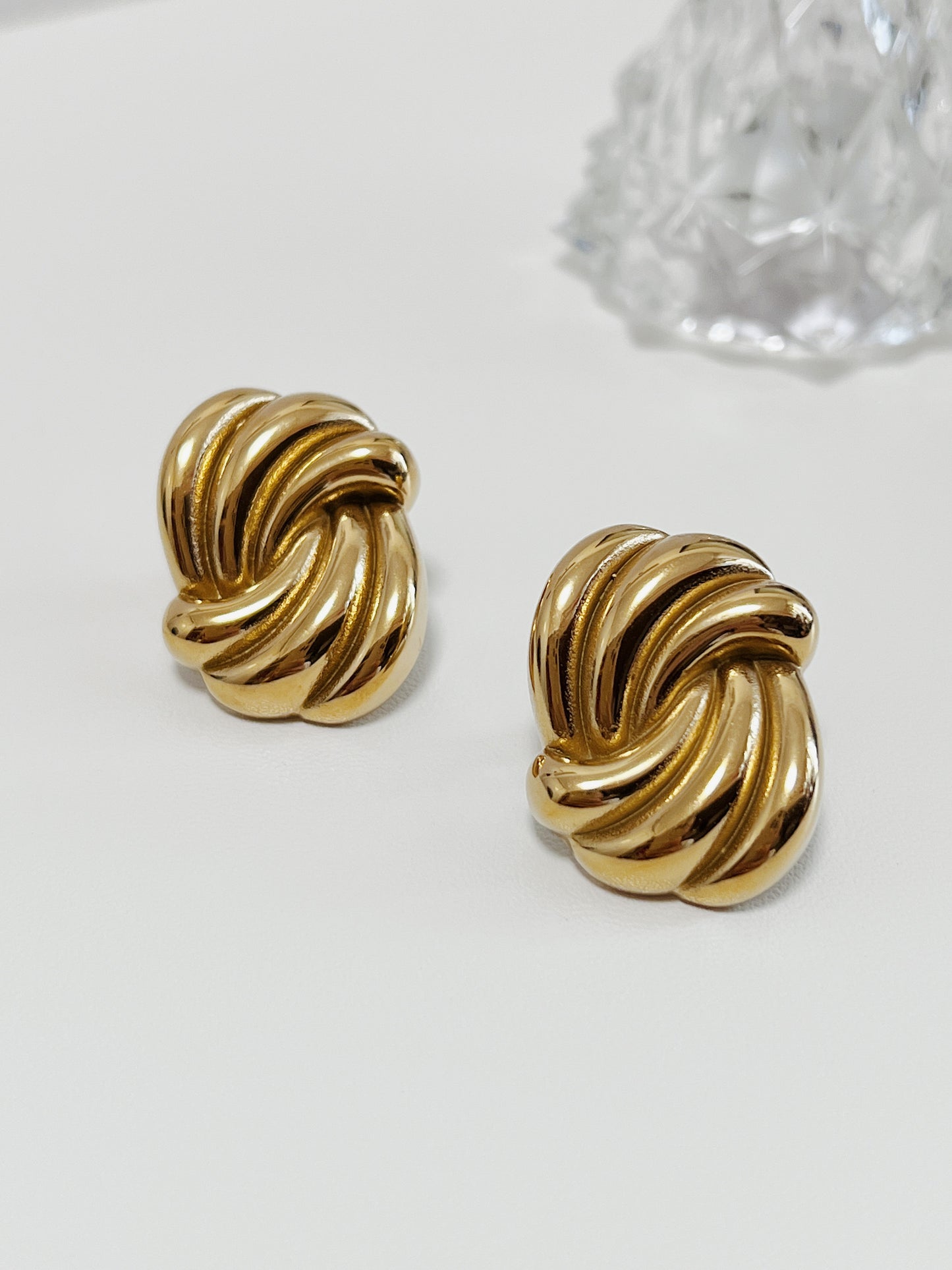Pair of gold twist knot earrings on a white surface with a crystal object in the background.