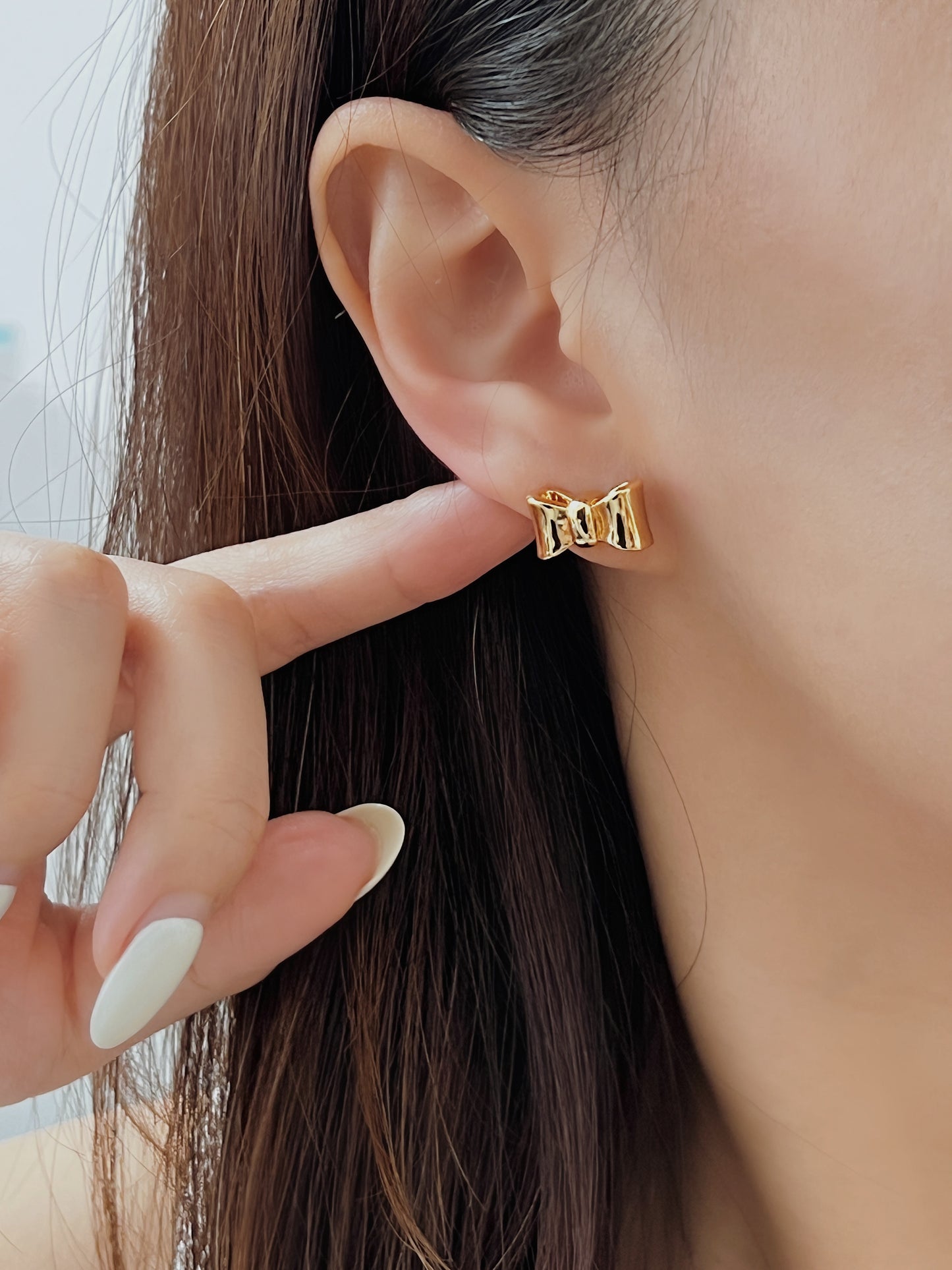 Gold bow-shaped earring being pointed at by a hand with light nail polish.