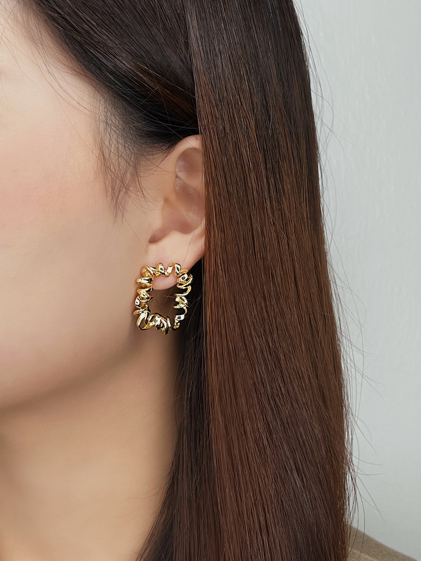 Gold curly earrings worn by a person with long brown hair against a plain background