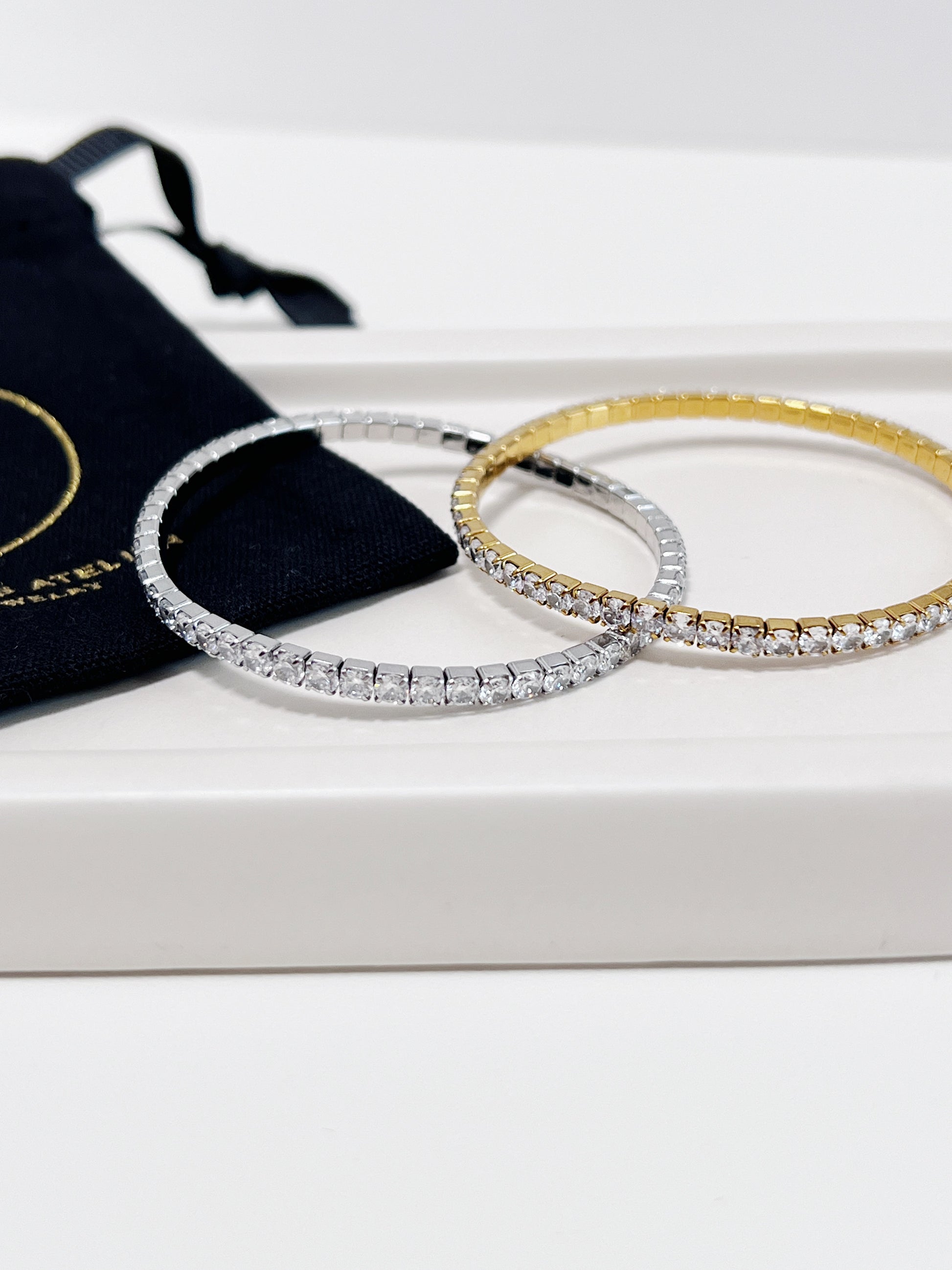 Two diamond tennis bracelets, one gold and one silver, on a beige ceramic plate with white surface and a black pouch.