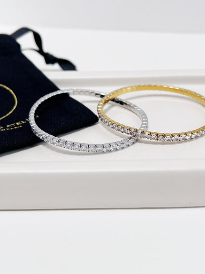 Two diamond tennis bracelets, one gold and one silver, on a beige ceramic plate with white surface and a black pouch.