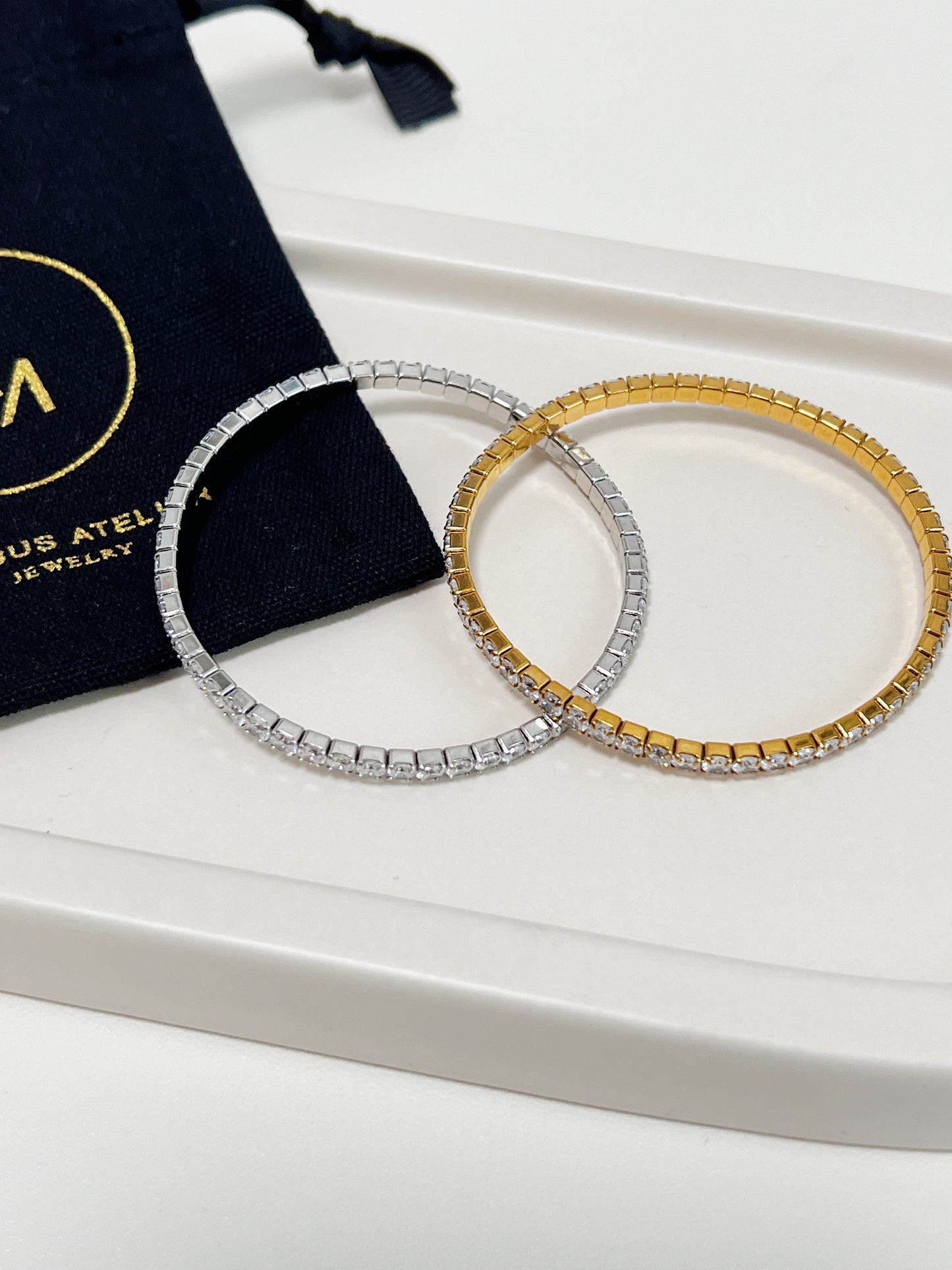 Two bracelets, one silver and one gold, on a ceramic plate placed on white surface with a black pouch in the background.