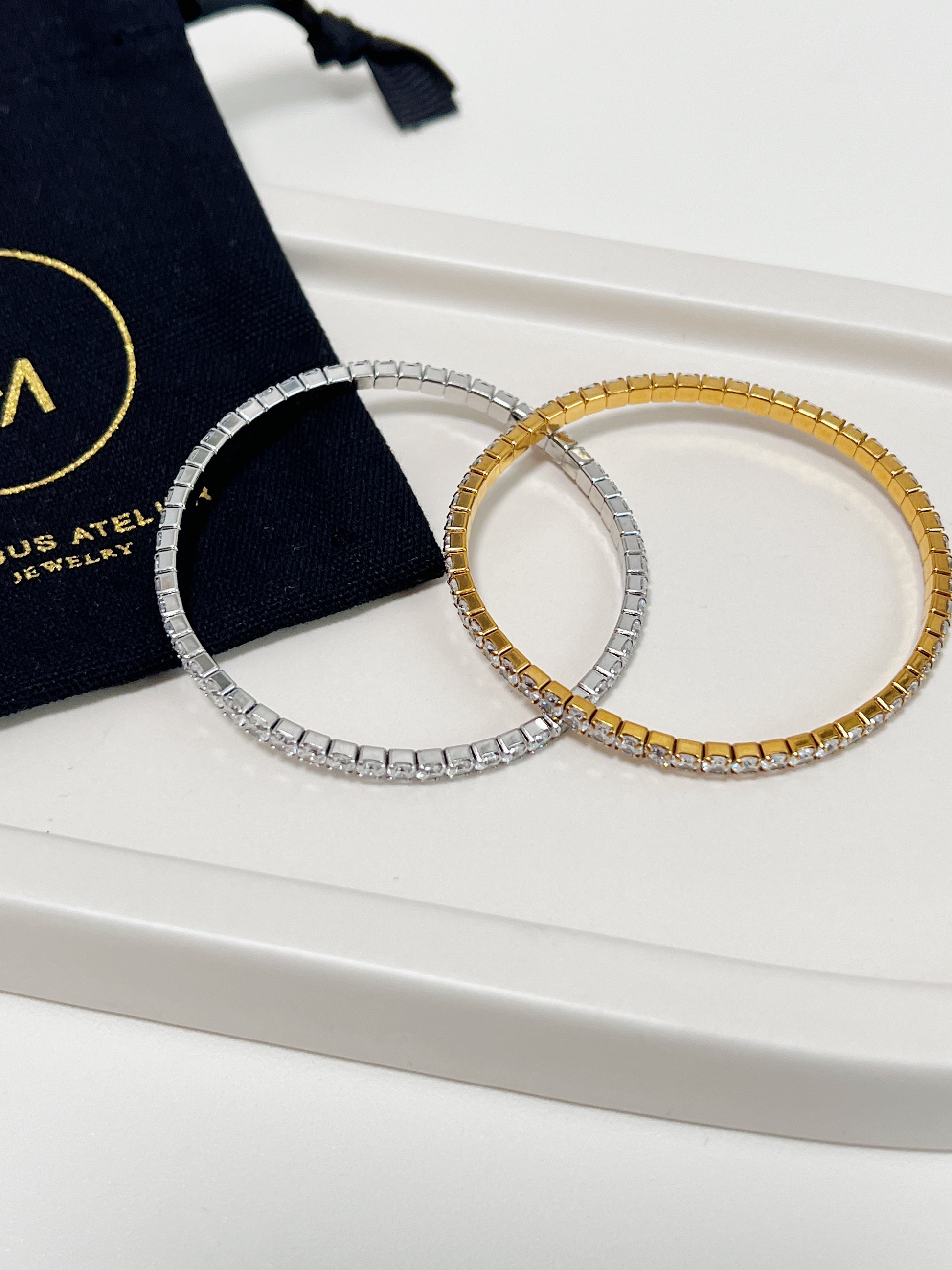 Two bracelets, one silver and one gold, on a ceramic plate placed on white surface with a black pouch in the background.