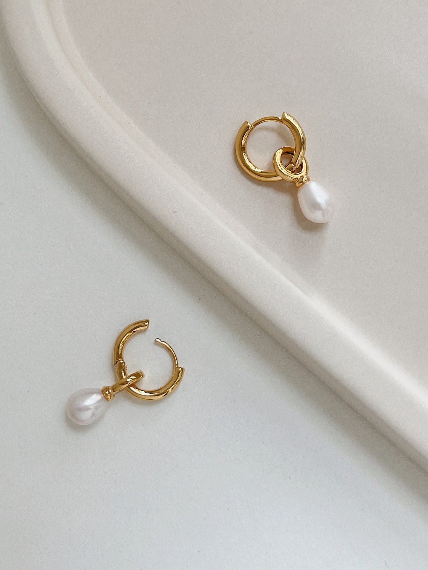 A pair of gold dangle pearl earrings, one placing on top of a white table and another one placing on top of a beige tray plate.