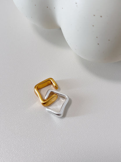 Gold and silver square-shaped ear cuff on a light gray surface with a white ceramic object in the background.