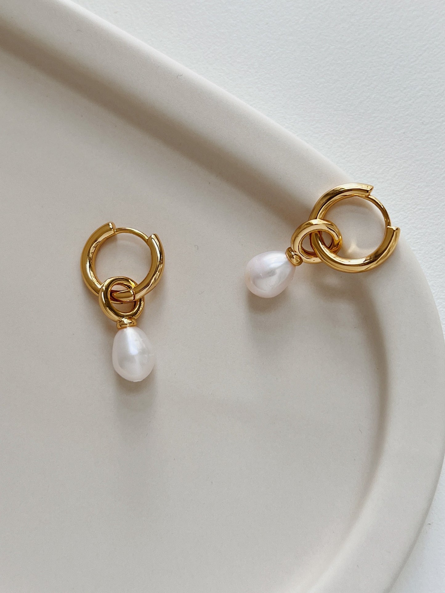 A pair of gold dangle pearl earrings placing on top of a beige tray plate.