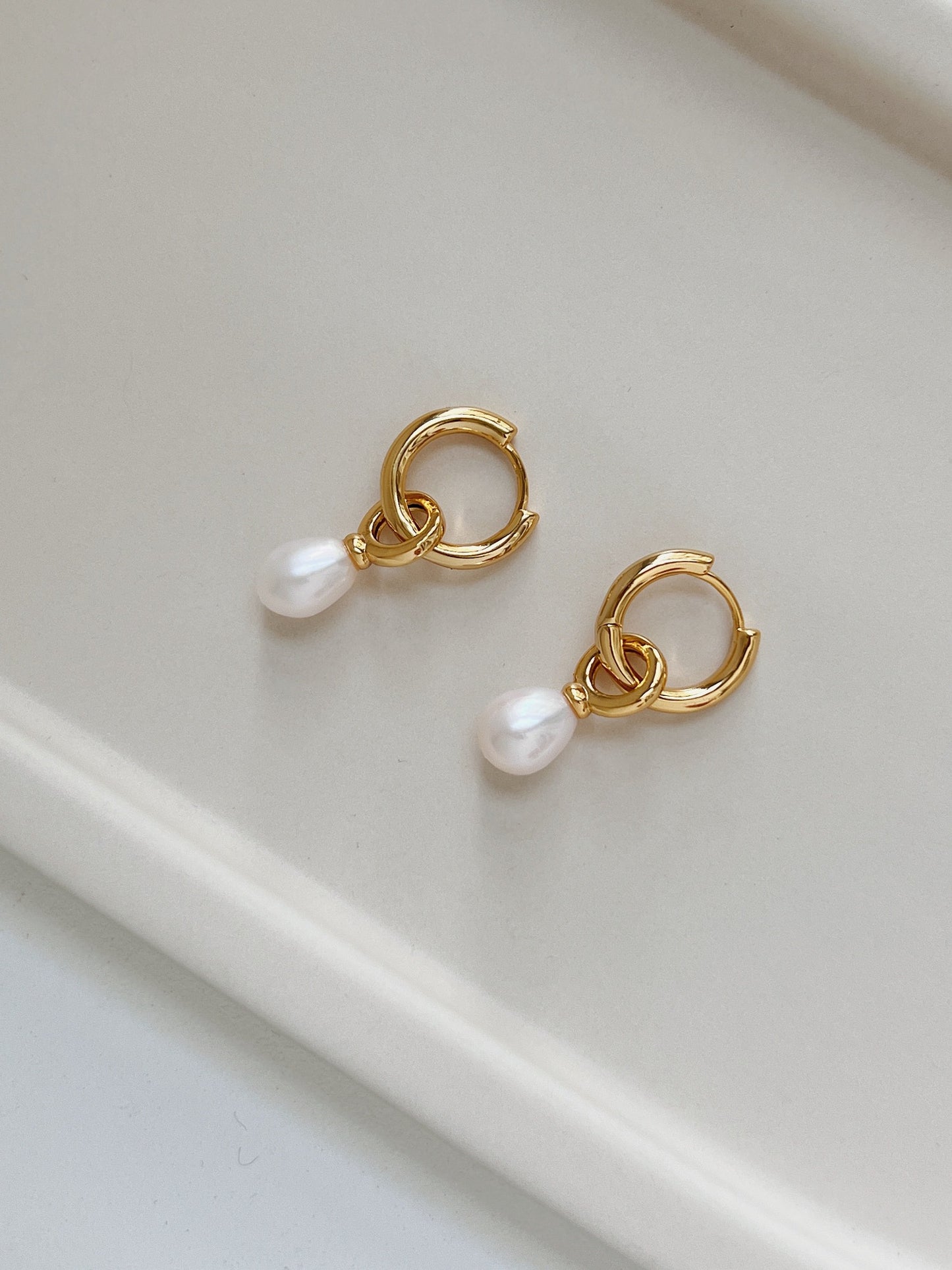 A pair of gold dangle pearl earrings placing on top of a beige tray plate.