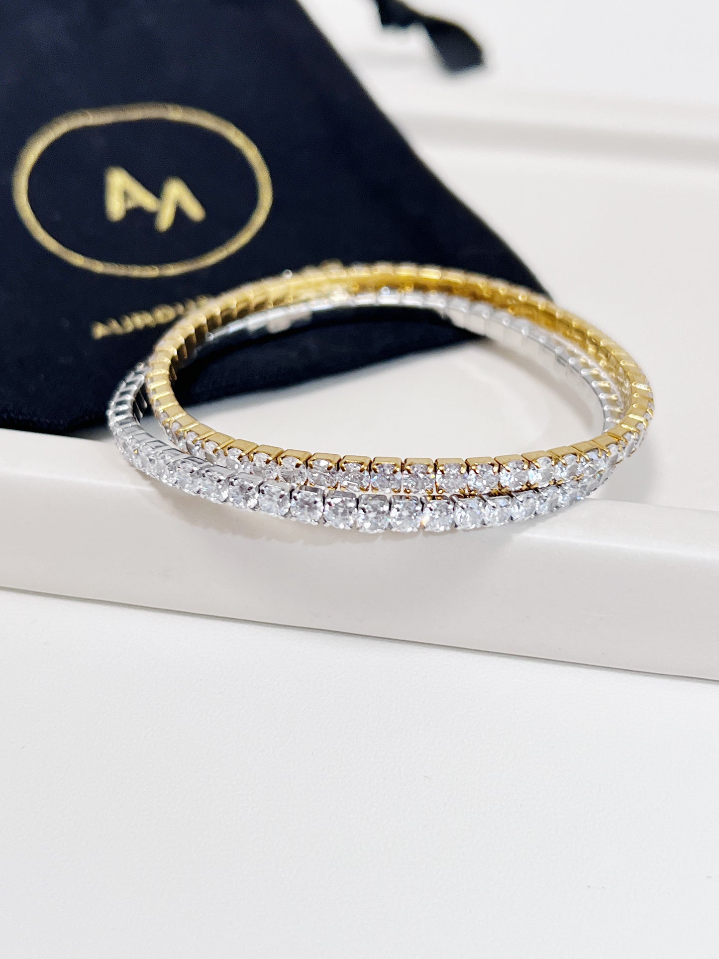 Gold and silver elastic tennis bracelet on a white surface with a black pouch in the background