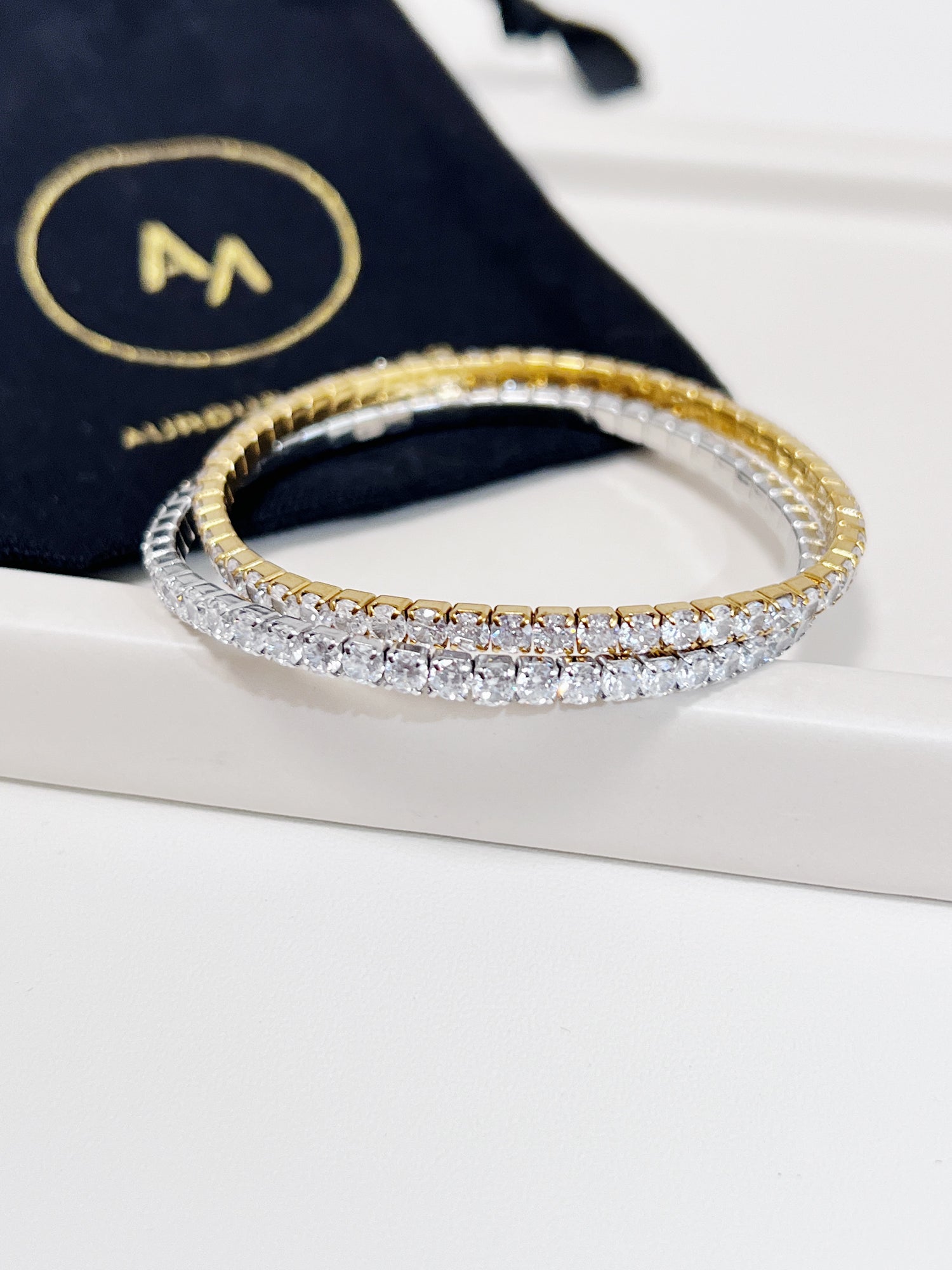 Gold and silver elastic tennis bracelet on a white surface with a black pouch in the background