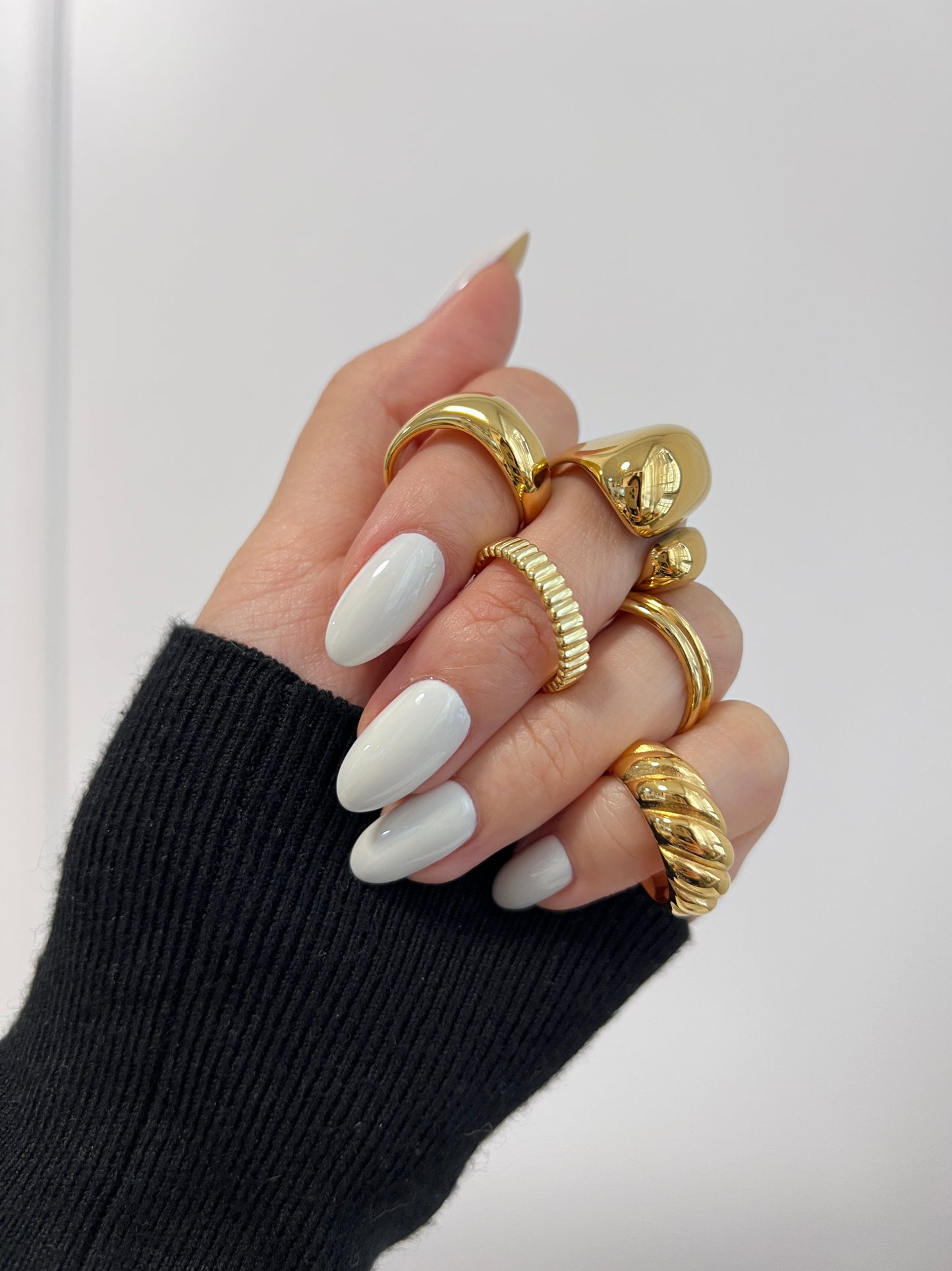 Hand wearing multiple gold rings with white nail polish on a light gray background