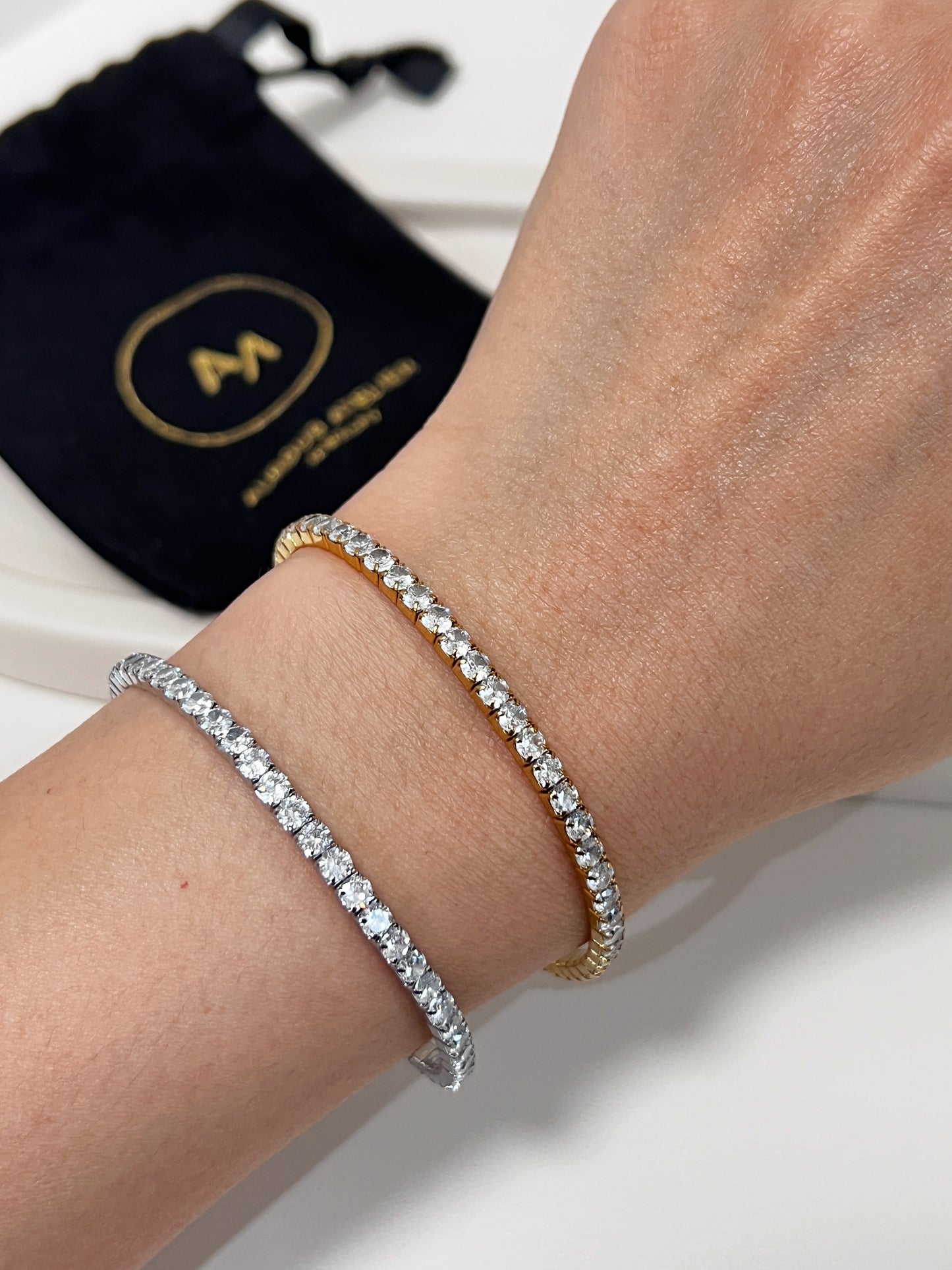 Two diamond bracelets on a wrist with a black pouch featuring 2 gold 'A' logo in the background.