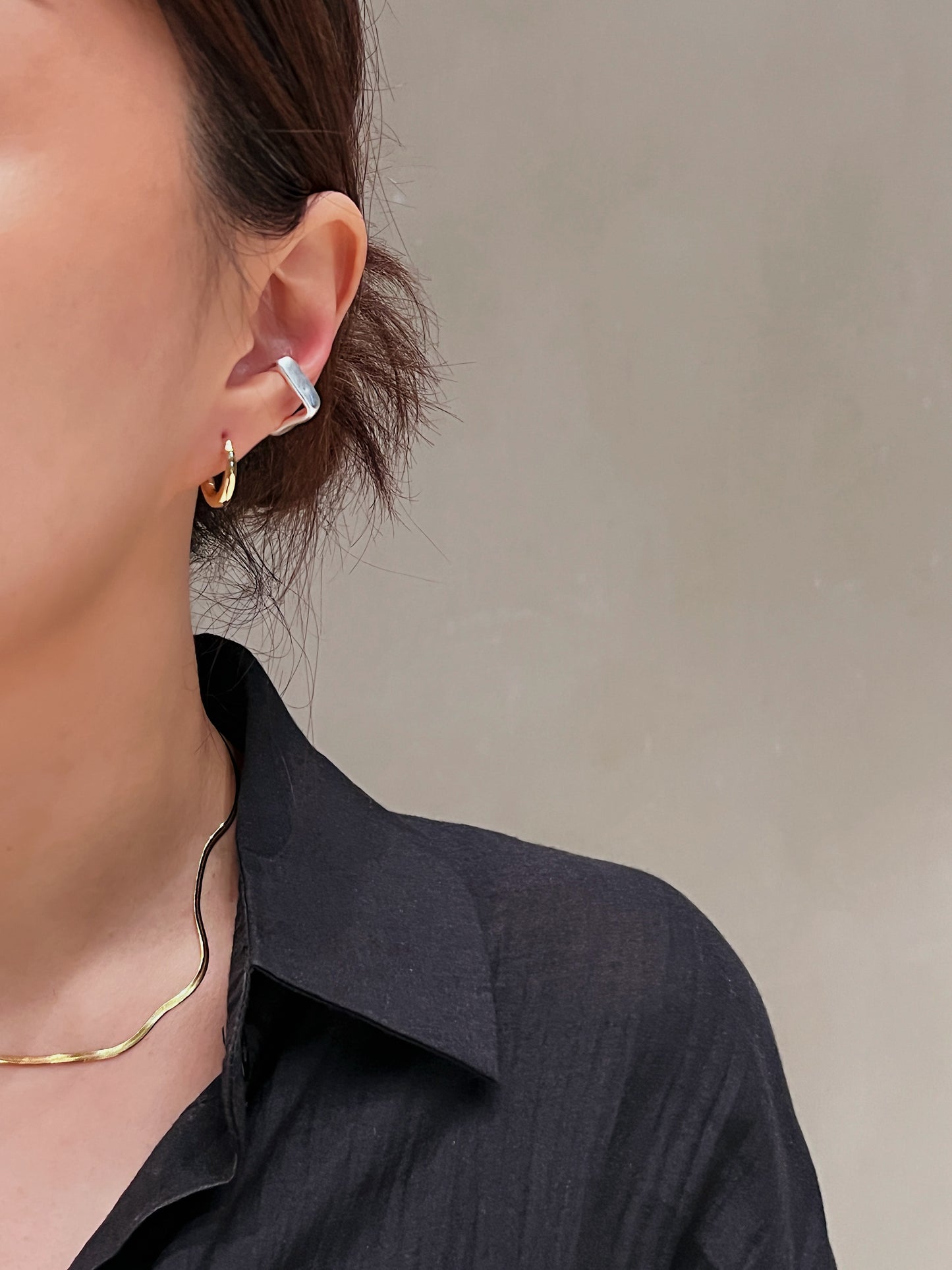 Close-up of a person wearing gold oval huggie hoop earrings and silver ear cuff with a neutral background