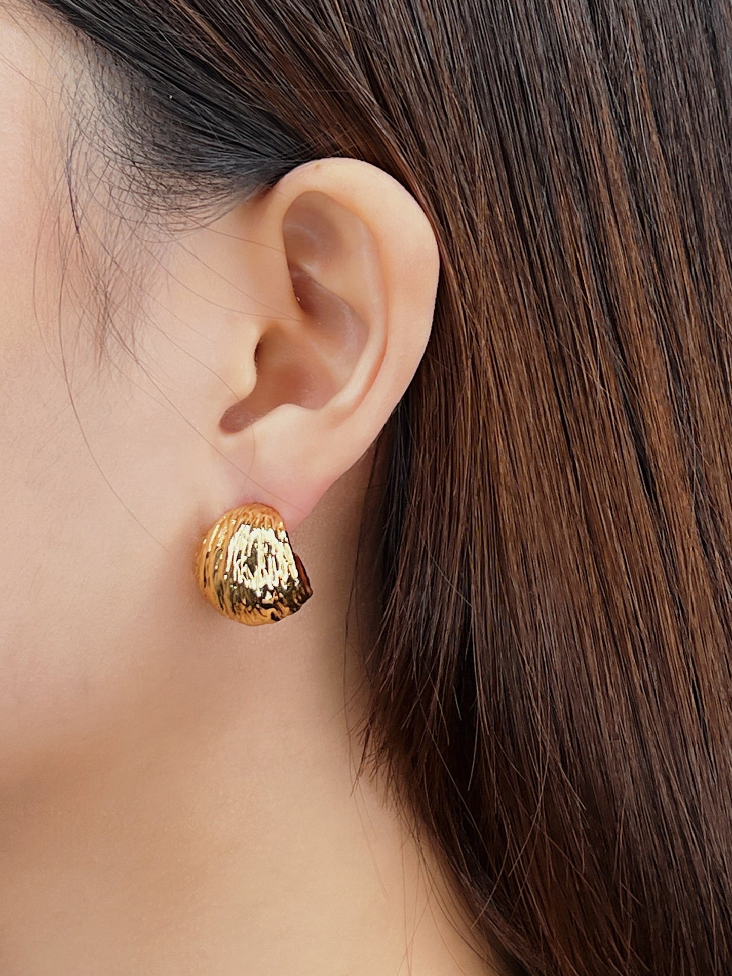 Gold textured earring worn by a person with long brown hair on a neutral background