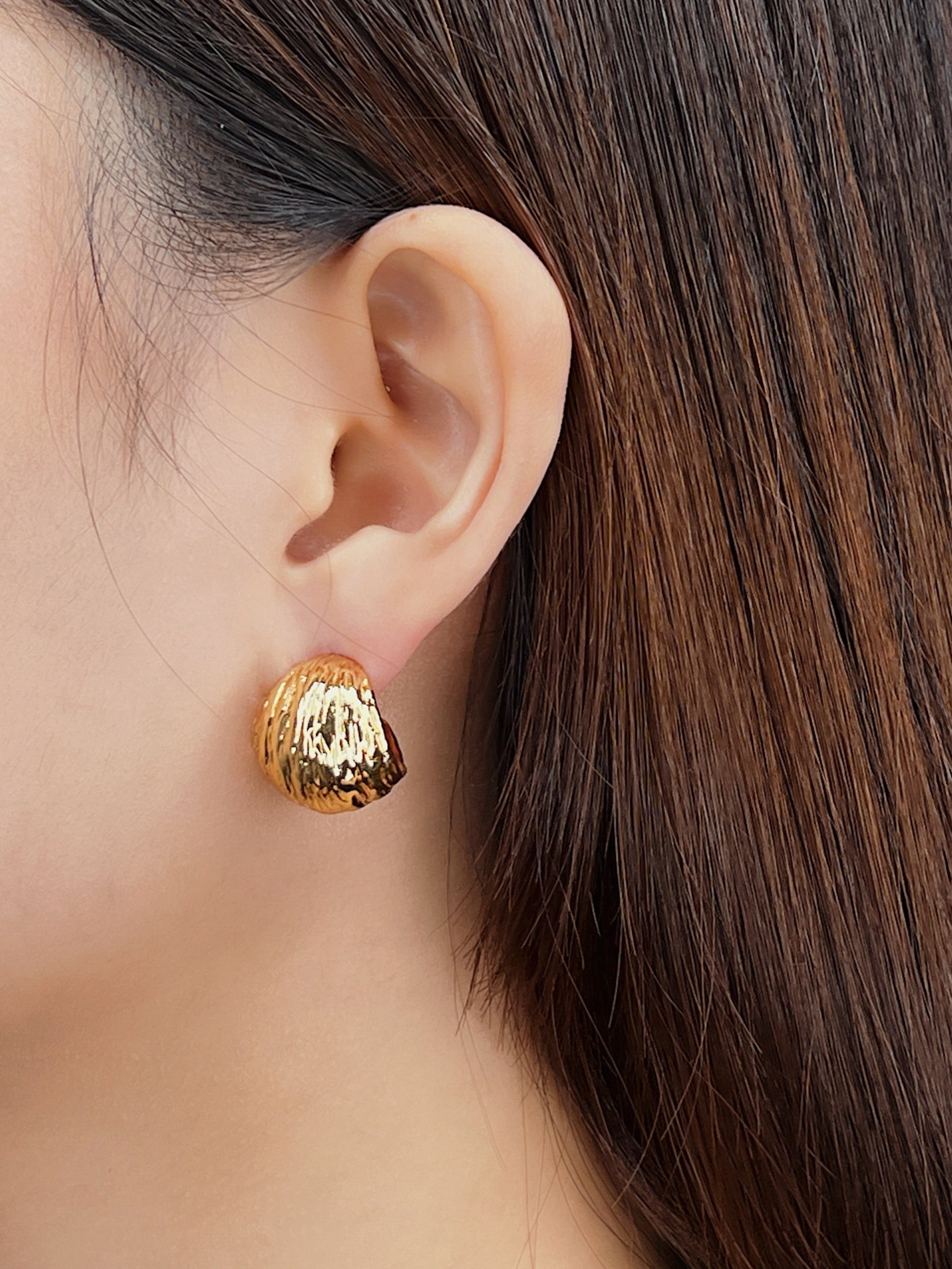Gold textured earring worn by a person with long brown hair on a neutral background