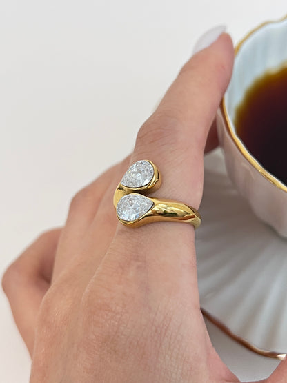 Gold ring with teardrop-shaped gemstones worn on a finger, with a blurred background of a cup of coffee.