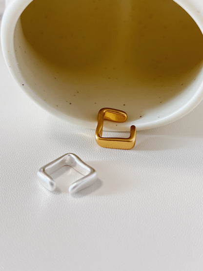 Gold and silver square-shaped ear cuff earrings on a white surface with a beige bowl in the background.