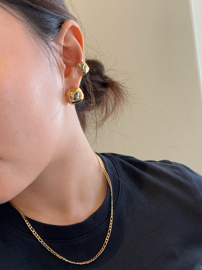 Close-up of a person wearing gold earrings and a gold figaro chain necklace against a neutral background