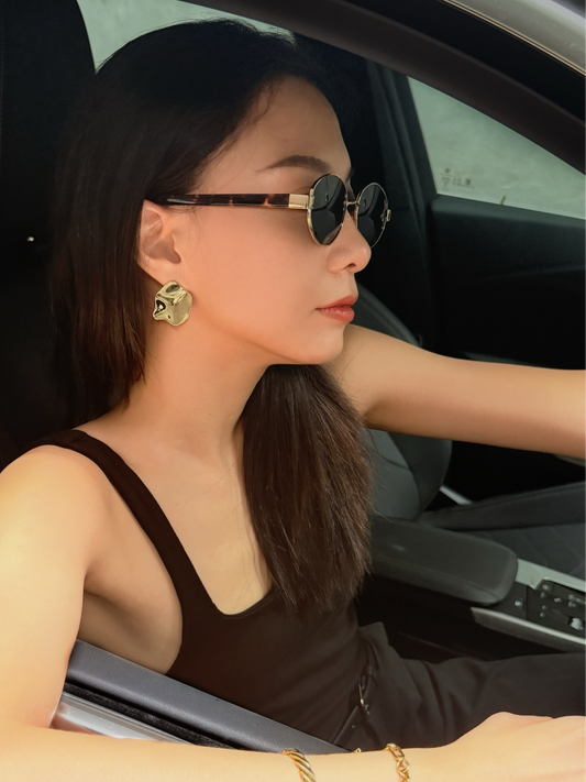 Woman sitting in a car wearing sunglasses, gold statement earrings and layering bracelets.