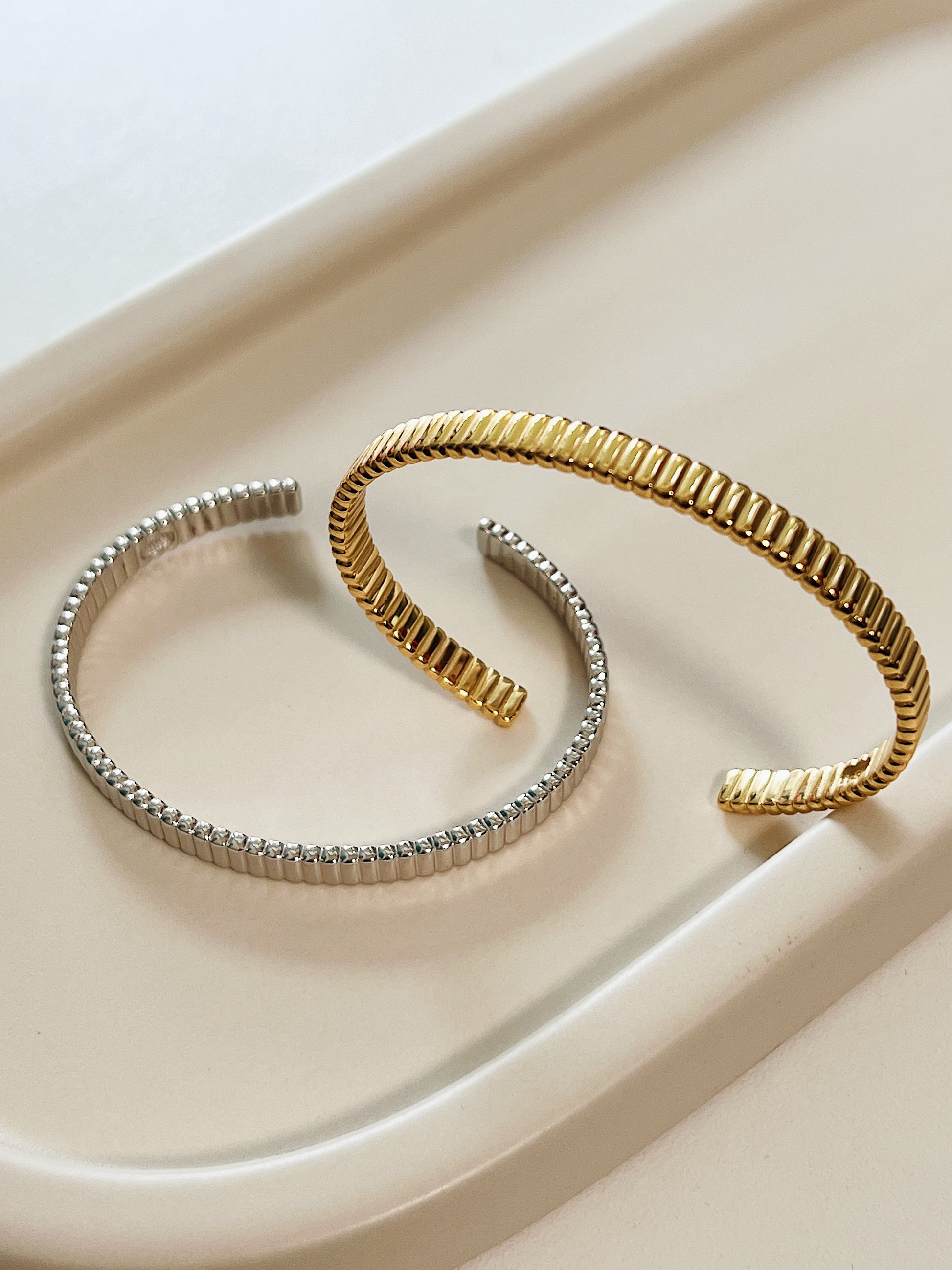 Overhead studio shot of the gold and silver adjustable open cuff bangles overlapping on a matte surface to showcase their flexible, structural columns.