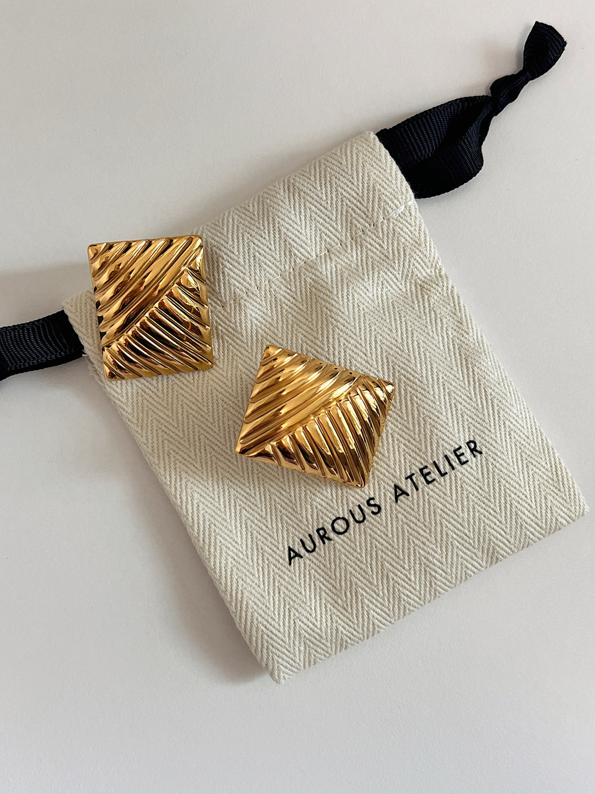 Pair of 18k gold pvd coated ribbed rectangle statement studs featuring dual-directional linear grooves and high-shine mirror polish resting on a cream canvas Aurous Atelier pouch