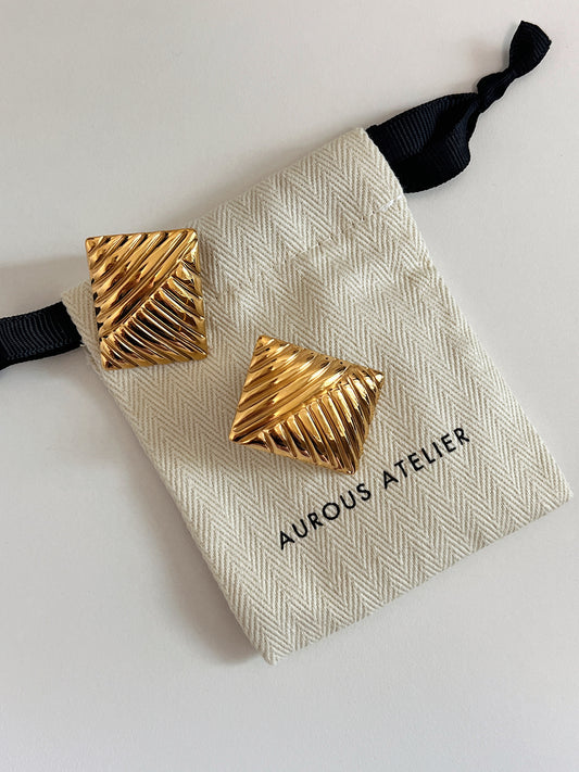 Pair of 18k gold pvd coated ribbed rectangle statement studs featuring dual-directional linear grooves and high-shine mirror polish resting on a cream canvas Aurous Atelier pouch