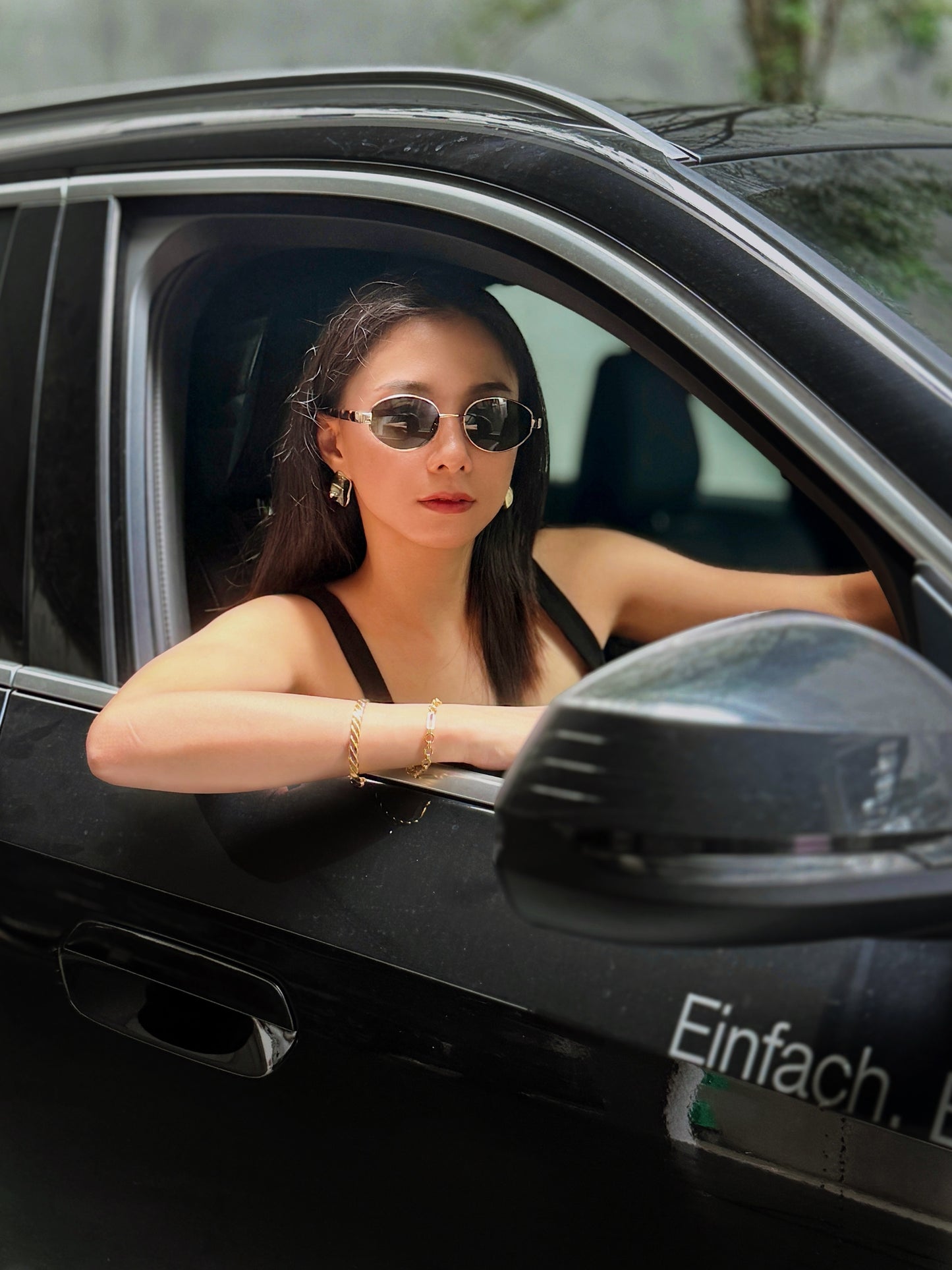 Model wearing Amalfi gold wire sunglasses while driving a car, showcasing the high-fashion look of the deep green lenses against a black outfit.