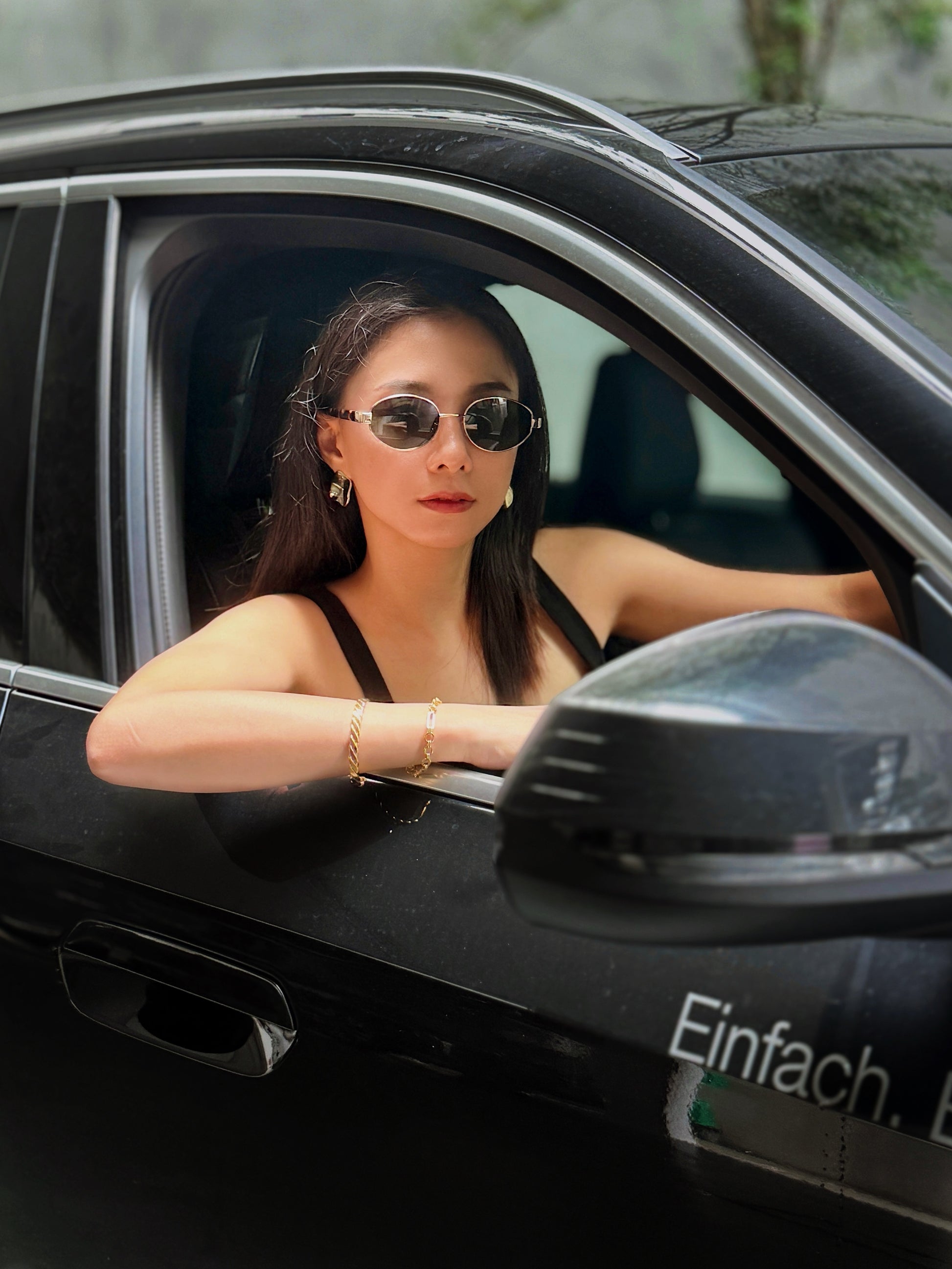 Model wearing Amalfi gold wire sunglasses while driving a car, showcasing the high-fashion look of the deep green lenses against a black outfit.