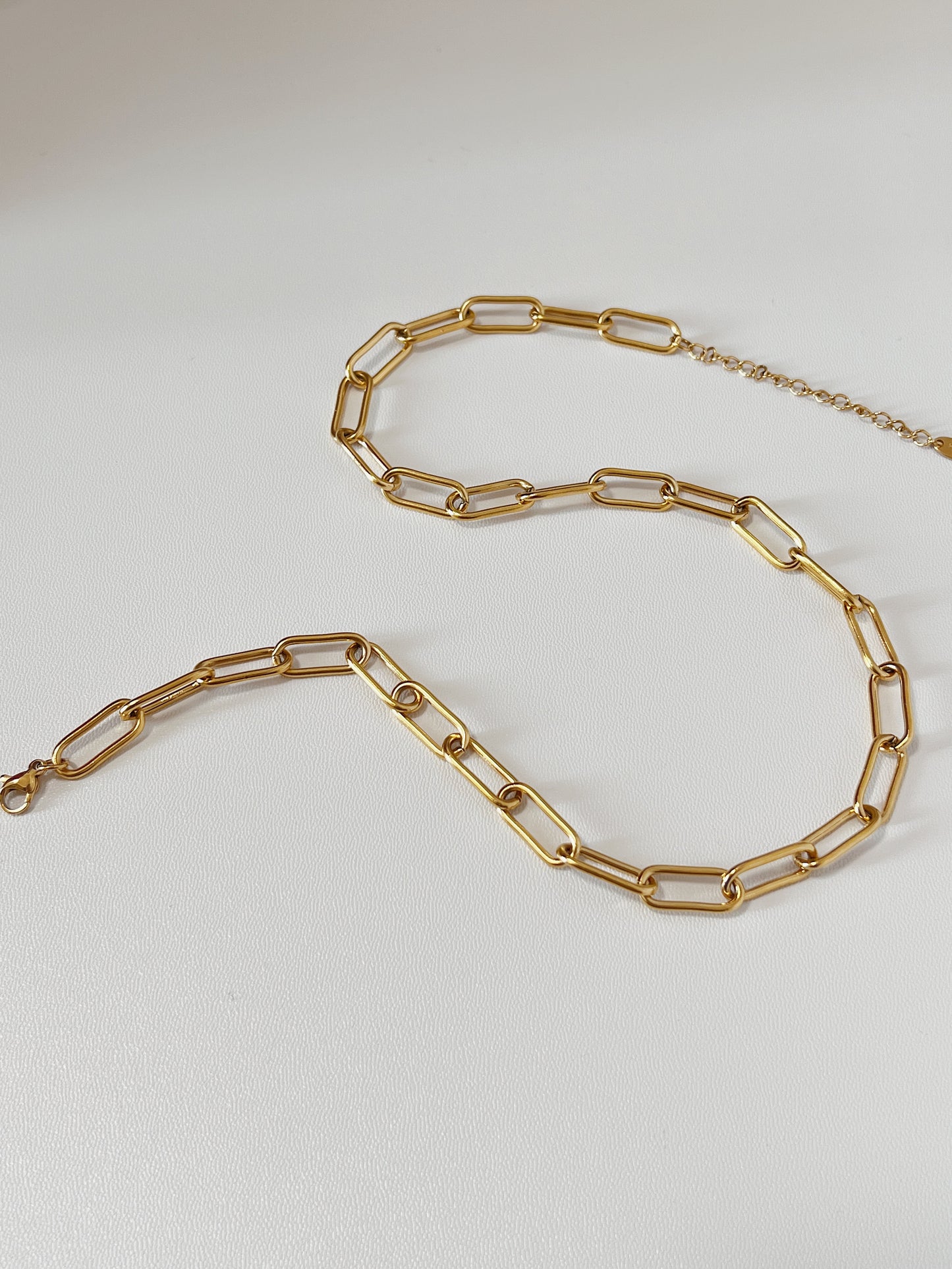 Close-up product photography of Asher chunky gold link necklace laid out in a fluid S-curve silhouette on a clean white surface, highlighting the geometric paperclip link design.