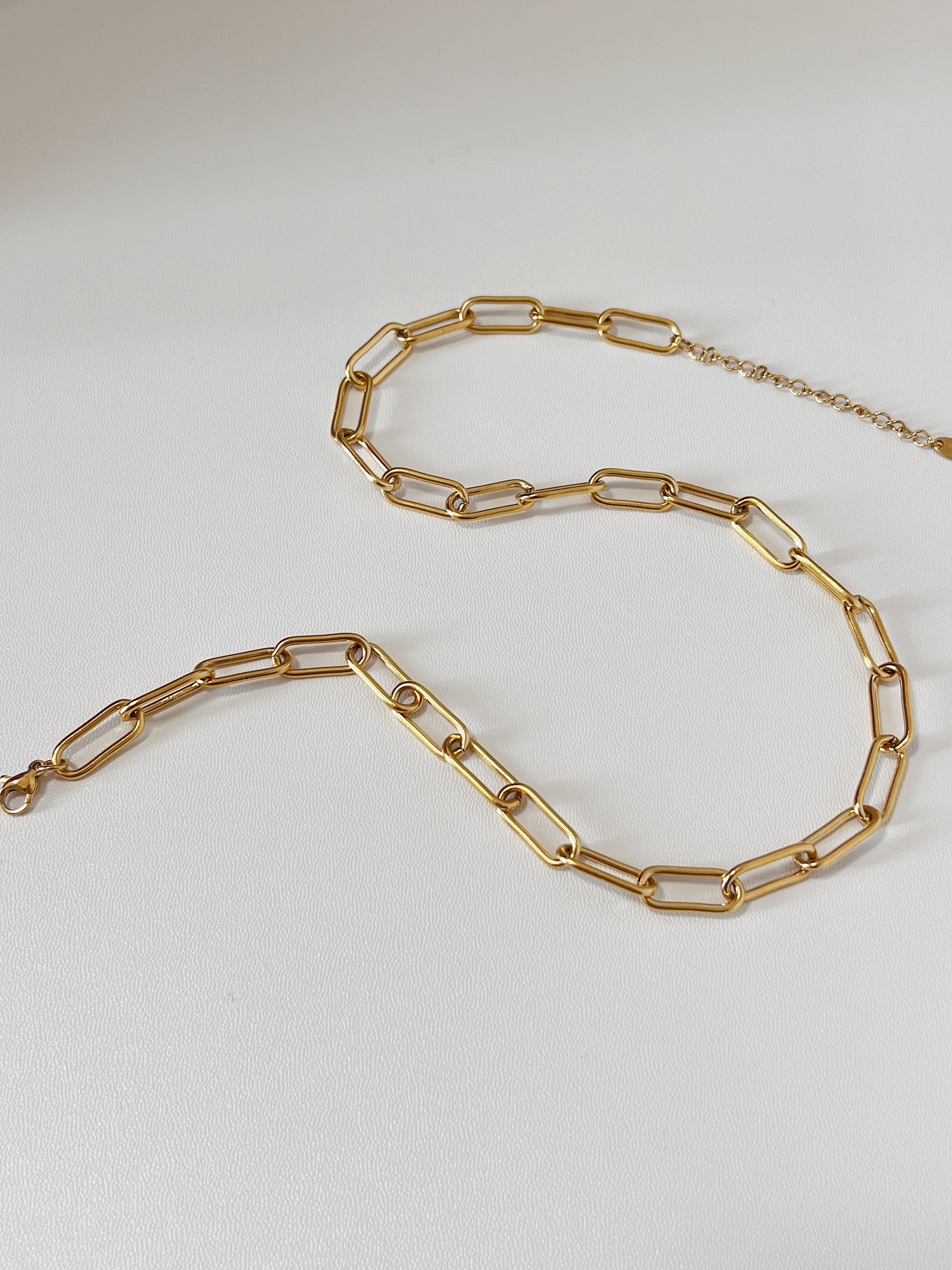 Close-up product photography of Asher chunky gold link necklace laid out in a fluid S-curve silhouette on a clean white surface, highlighting the geometric paperclip link design.