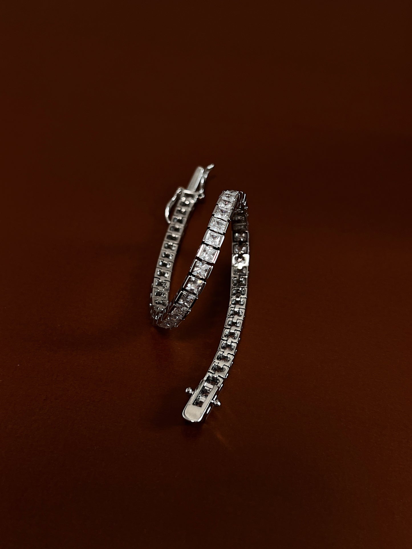 Coiled view displaying the fluid articulated link structure and edge-to-edge metallic channel holding 3mm square-cut cubic zirconia stones on a deep brown surface.