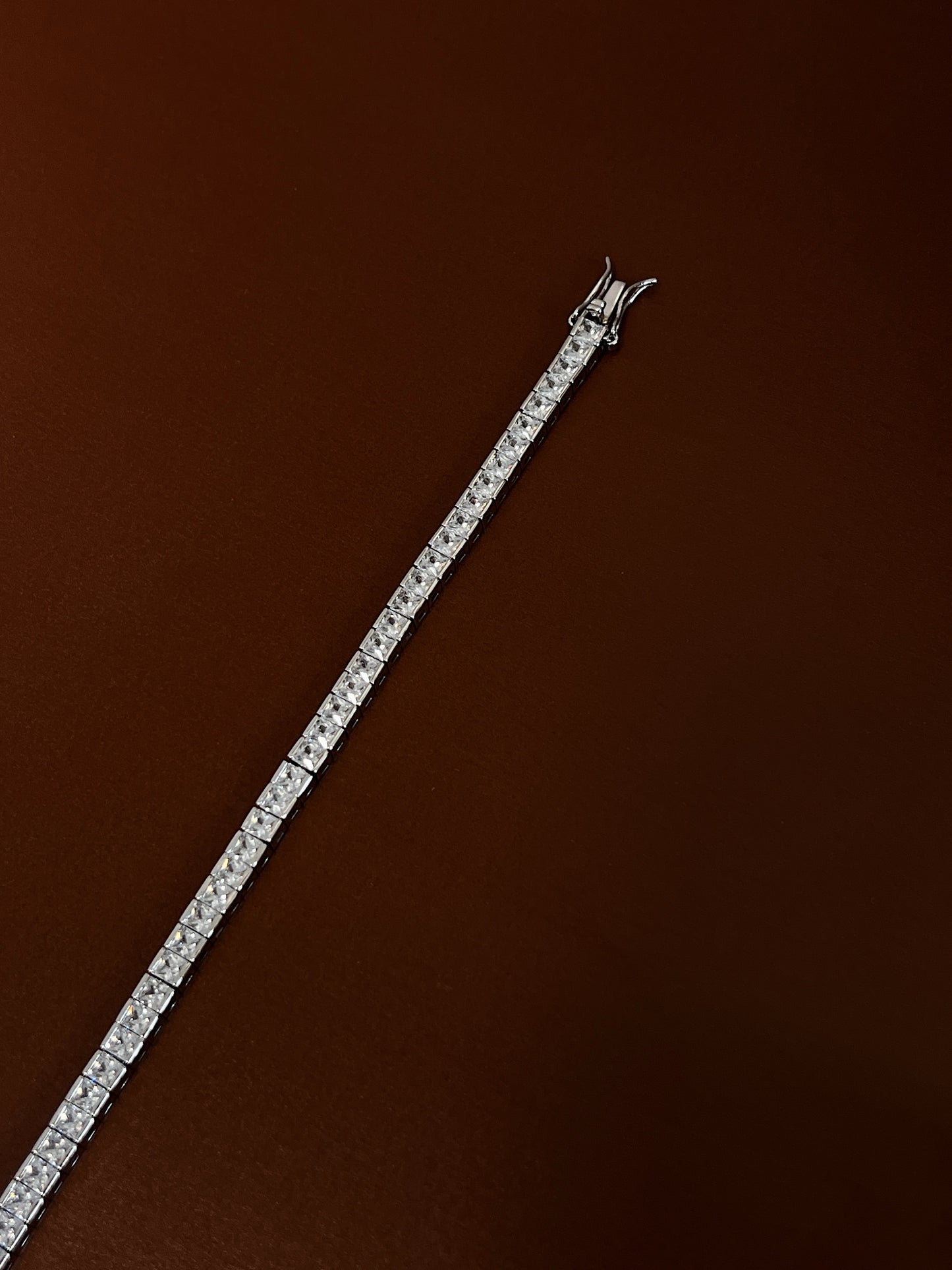 High-luster rhodium finish gleaming across the linear channel setting of a square-cut silver tennis bracelet on a dark background.