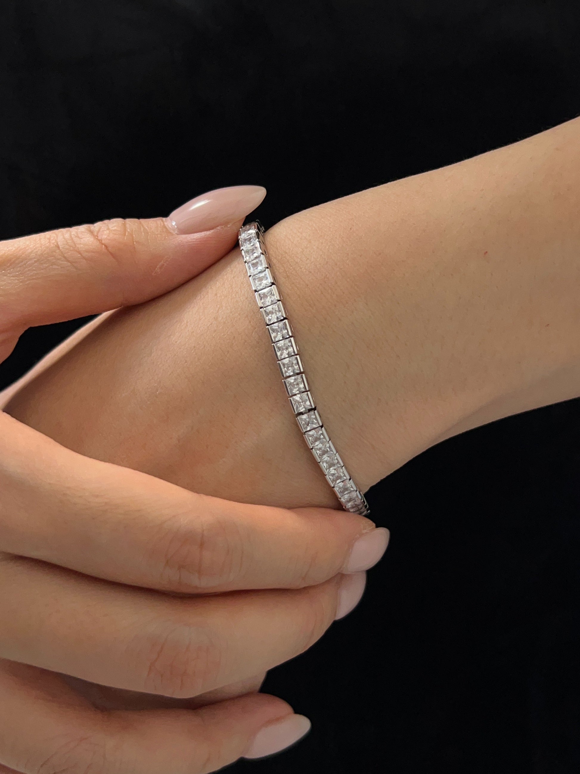 Close-up wrist view highlighting the high-shine rhodium finish and flush-mounted square stones resting against a clean nude manicure and dark background.