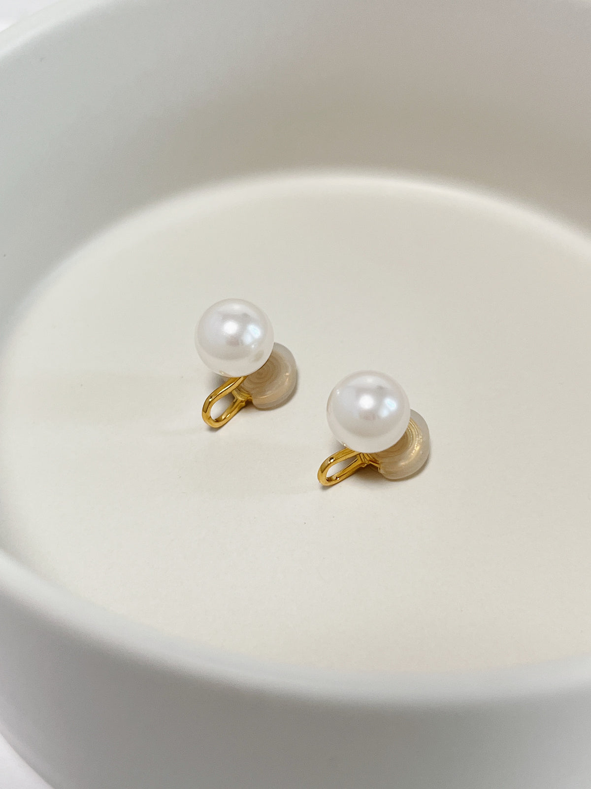 High-lustre 10mm pearl clip-on earrings resting on a white ceramic dish, showing 18k gold plated jeweler's brass wire and soft silicone pads.