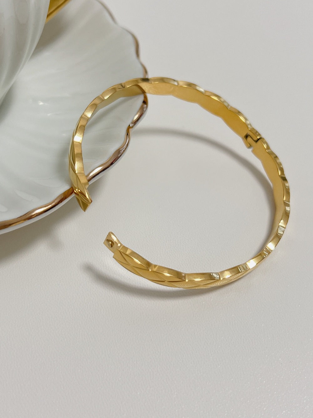 A close-up shot of the gold Corin quilted bangle showing its open high-security side hinge mechanism resting next to a white ceramic saucer.