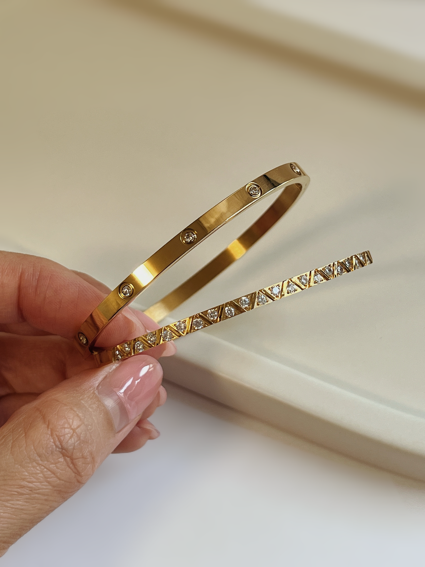 A close-up of a hand holding the Dane and Gia gold bangles, showcasing the polished gold finish, bezel-set stones, and geometric cubic zirconia details against a minimalist cream background.