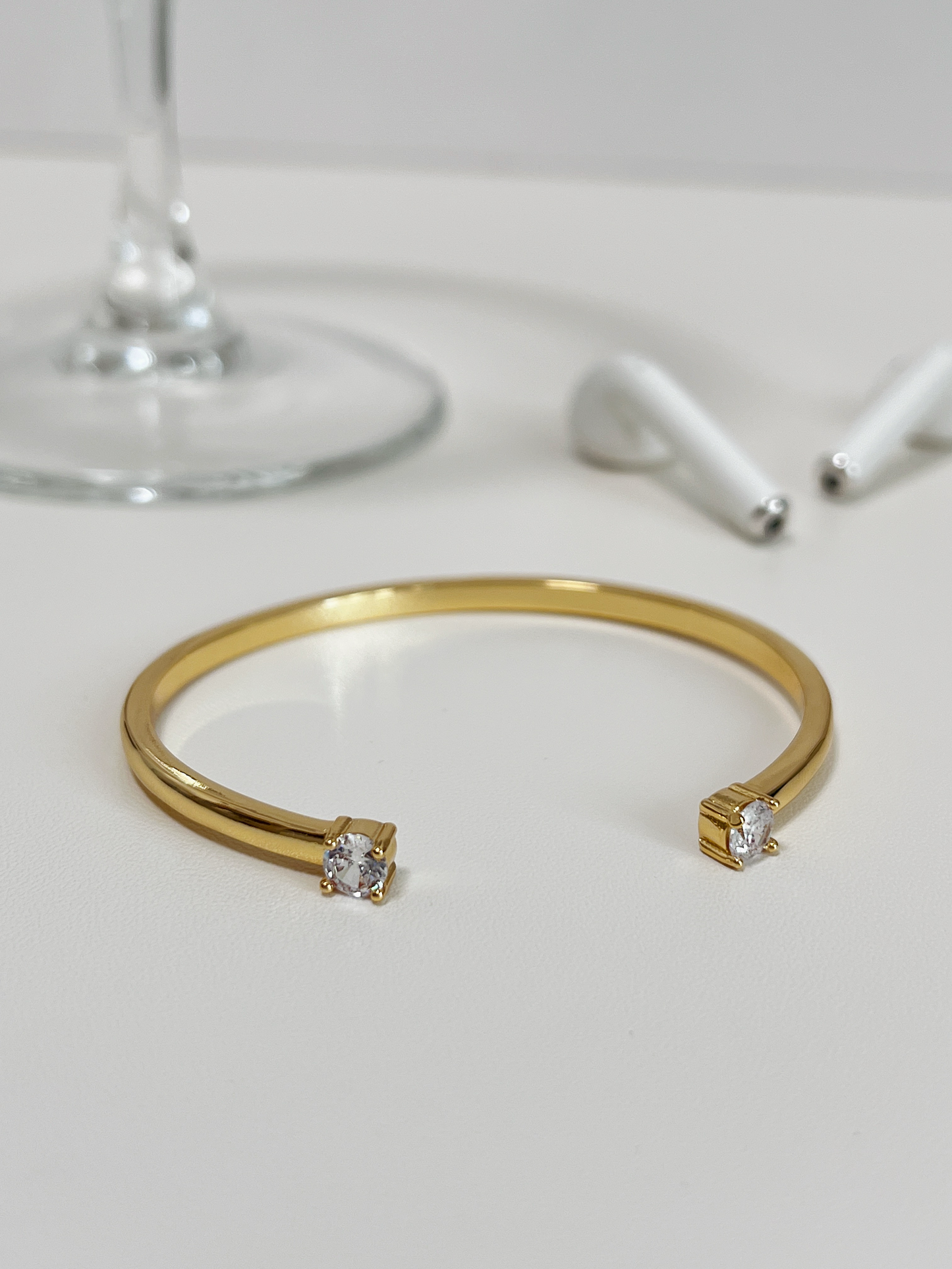 Minimalist product-focused flatlay of the Justine gold open-cuff bracelet resting on a clean white countertop next to wireless headphones.