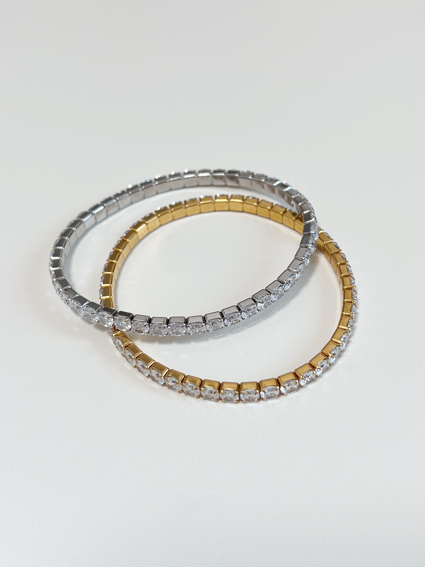 Overlapping gold and silver 4mm elastic tennis bracelets showcasing the smooth clasp-free stretch setting architecture.