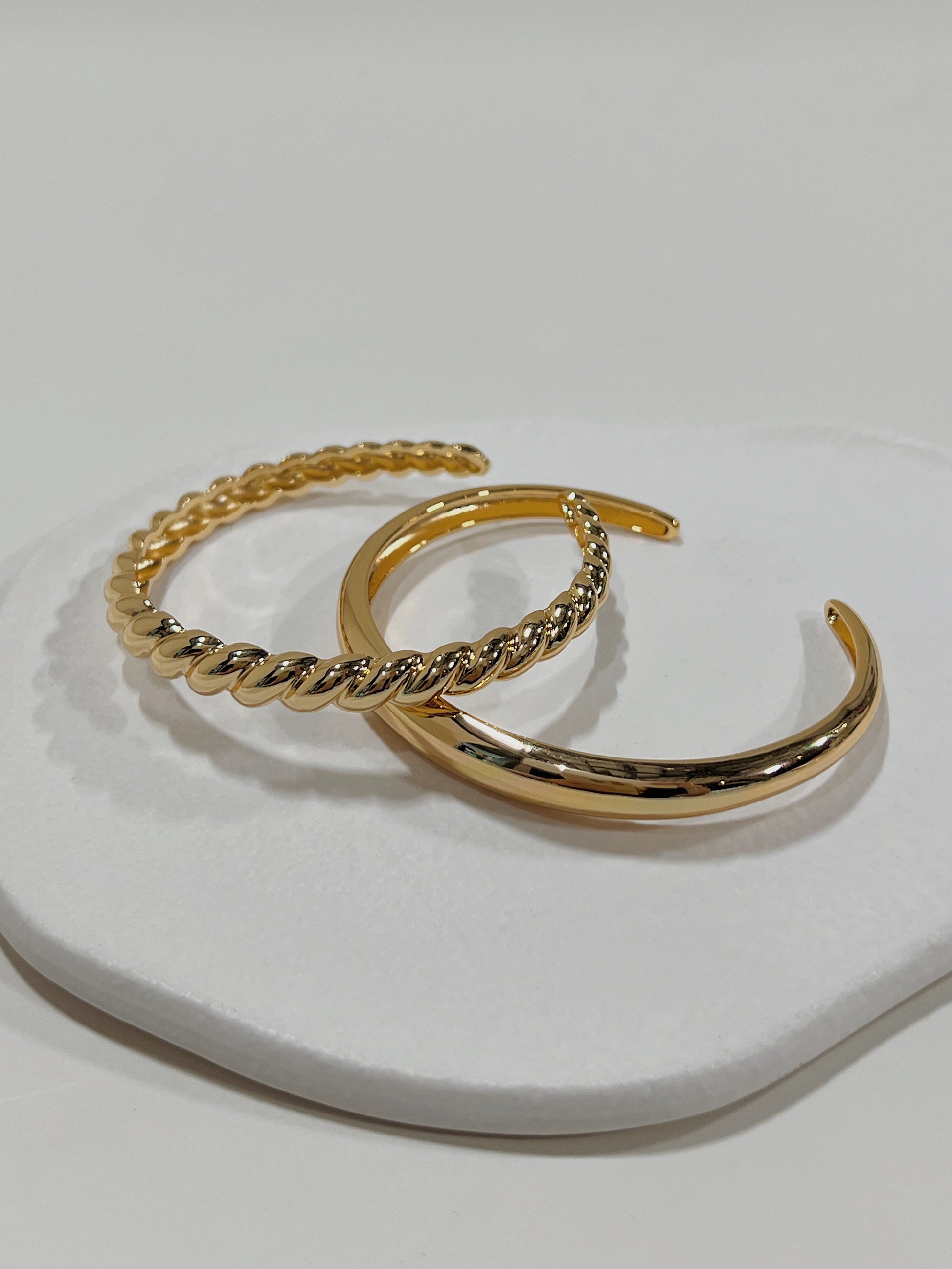 Architectural product shot featuring the Paris Croissant and London Plain gold cuff bracelets stacked together on a textured white background.