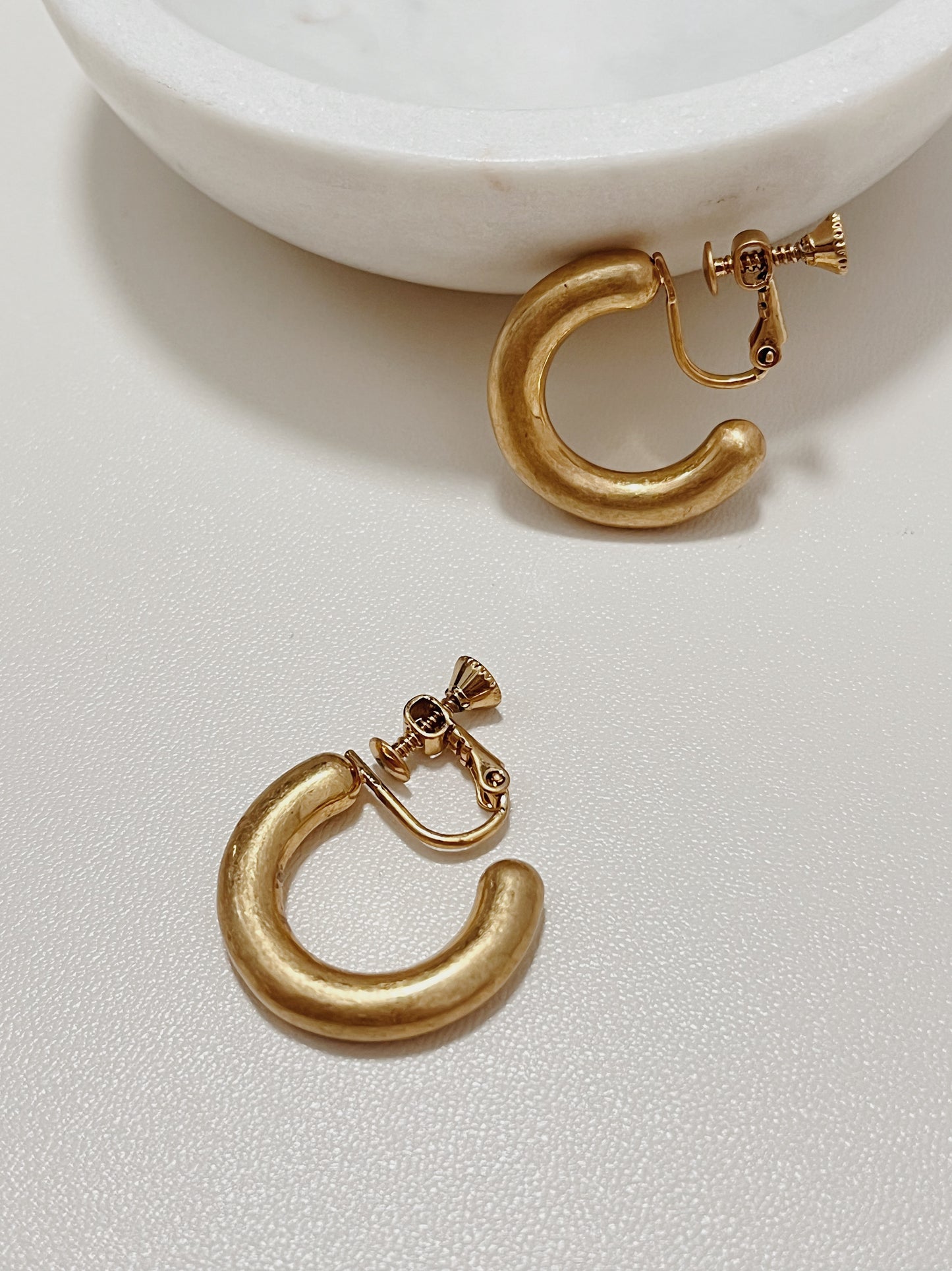 A pair of Zora chunky brushed gold clip-on hoop earrings with premium adjustable screw back clasps displayed on a white surface next to a minimal marble tray.
